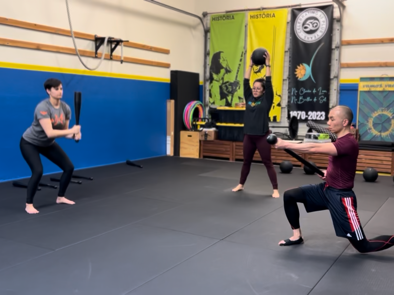 Three people working out in a gym. The woman in the center holds a medicine ball overhead, while the man on the right kneels with a weight plate, and the woman on the left holds a bat or club in a ready position.