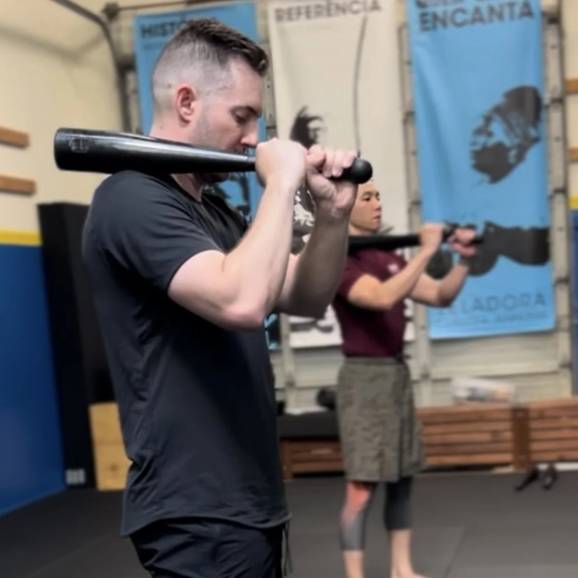 Two individuals are performing a workout with clubbells in a gym