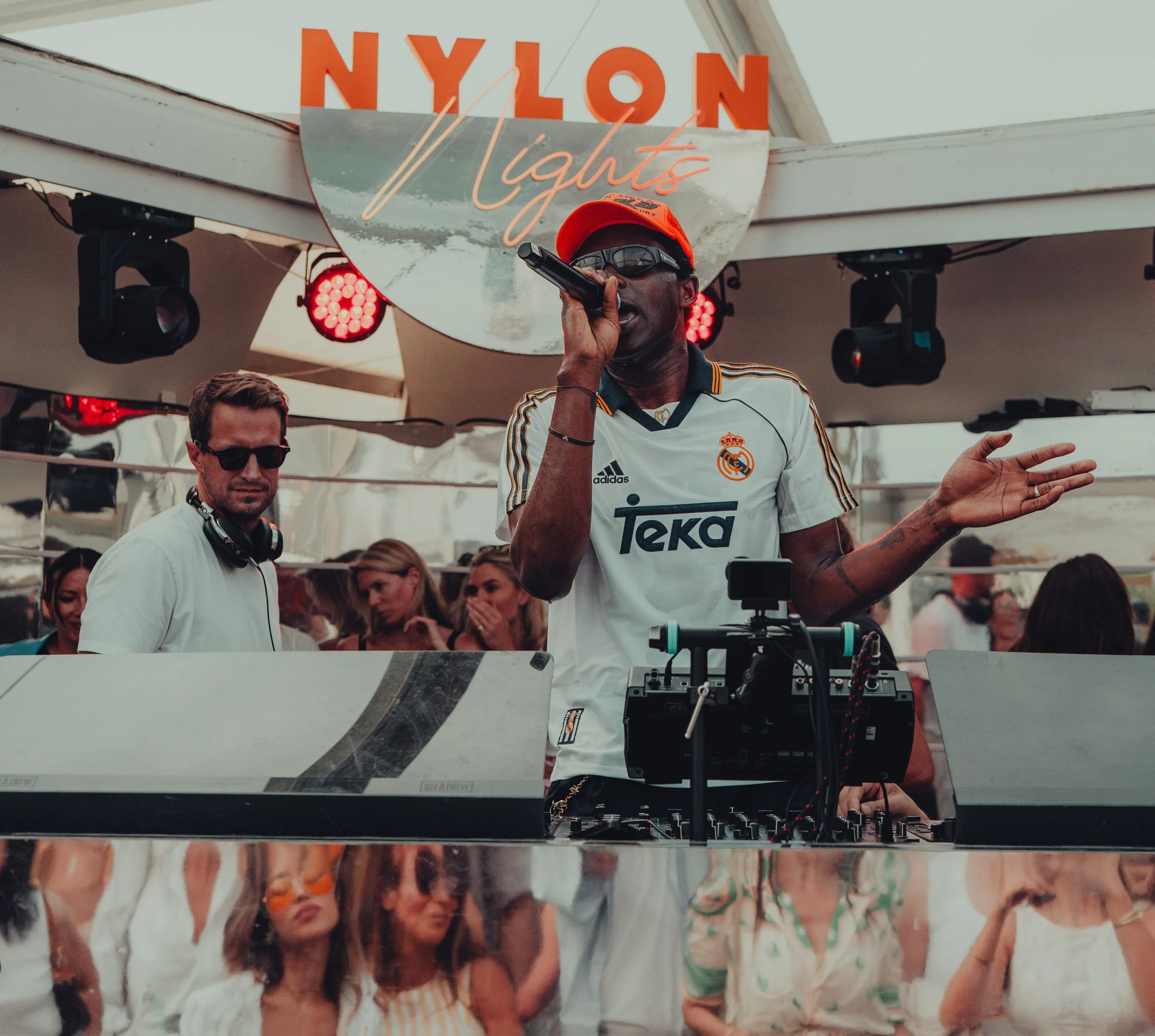 A DJ in a white Real Madrid jersey, red cap, and sunglasses is performing at an outdoor event under a canopy with a neon sign that reads 'NYLON Nights.' People are socializing and enjoying the music.