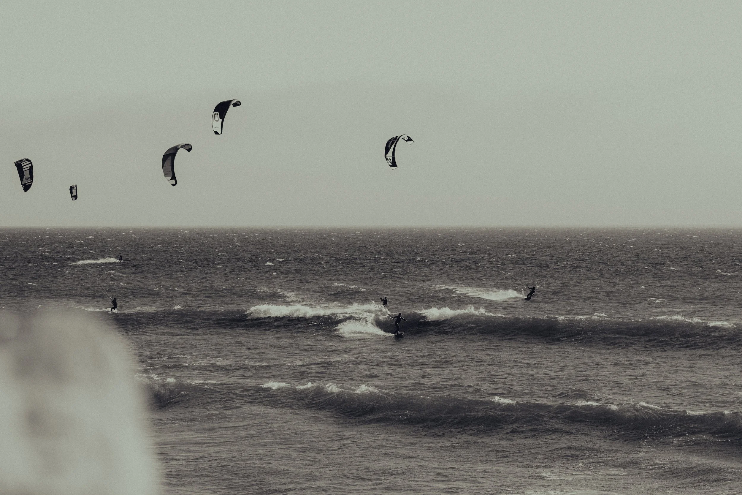 Multiple kite surfers riding waves in the ocean under a pale sky.