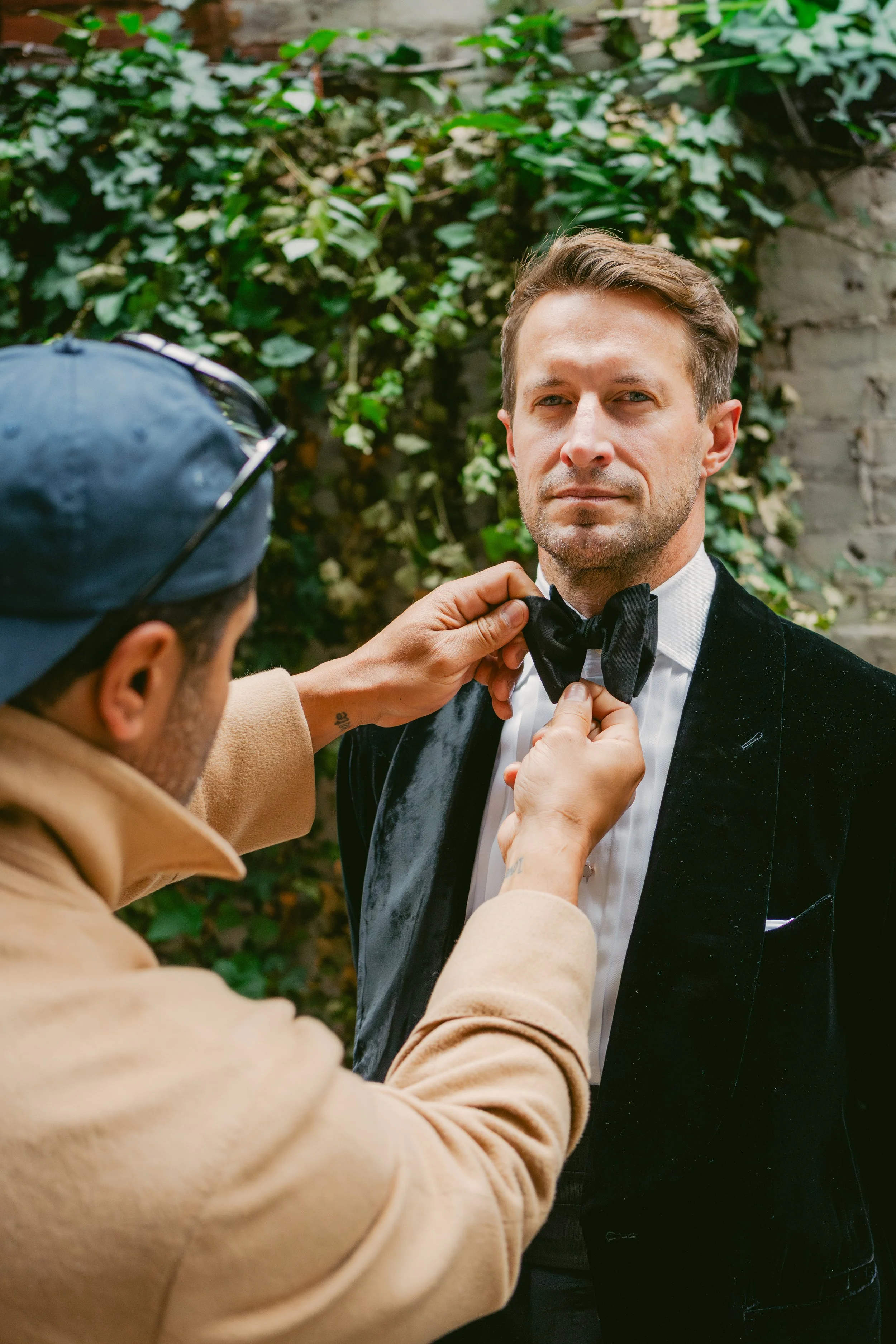 A man in a black tuxedo is being assisted with a black bowtie by another man in a beige coat and blue cap outdoors, with greenery in the background.