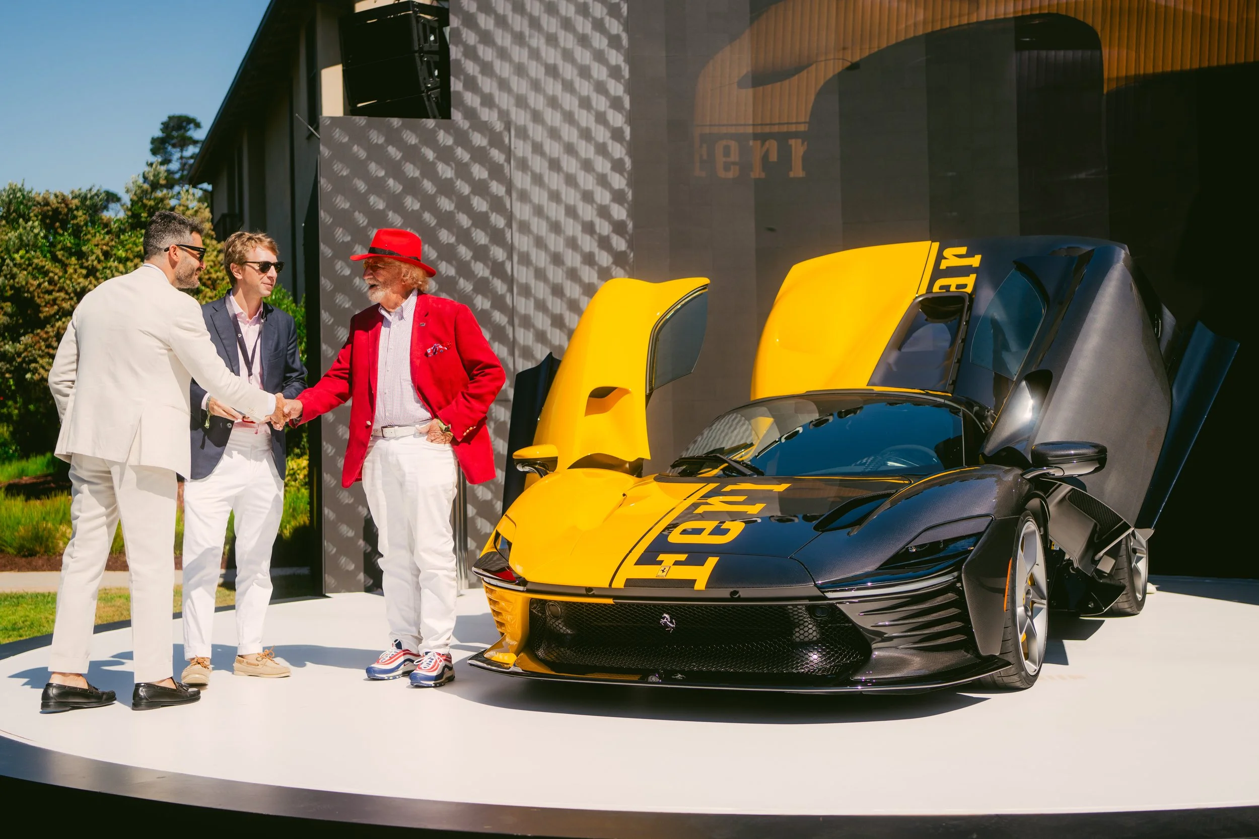 Four men dressed in stylish fashion, shaking hands next to a yellow and black Ferrari sports car with gullwing doors open, at an outdoor event on a sunny day.