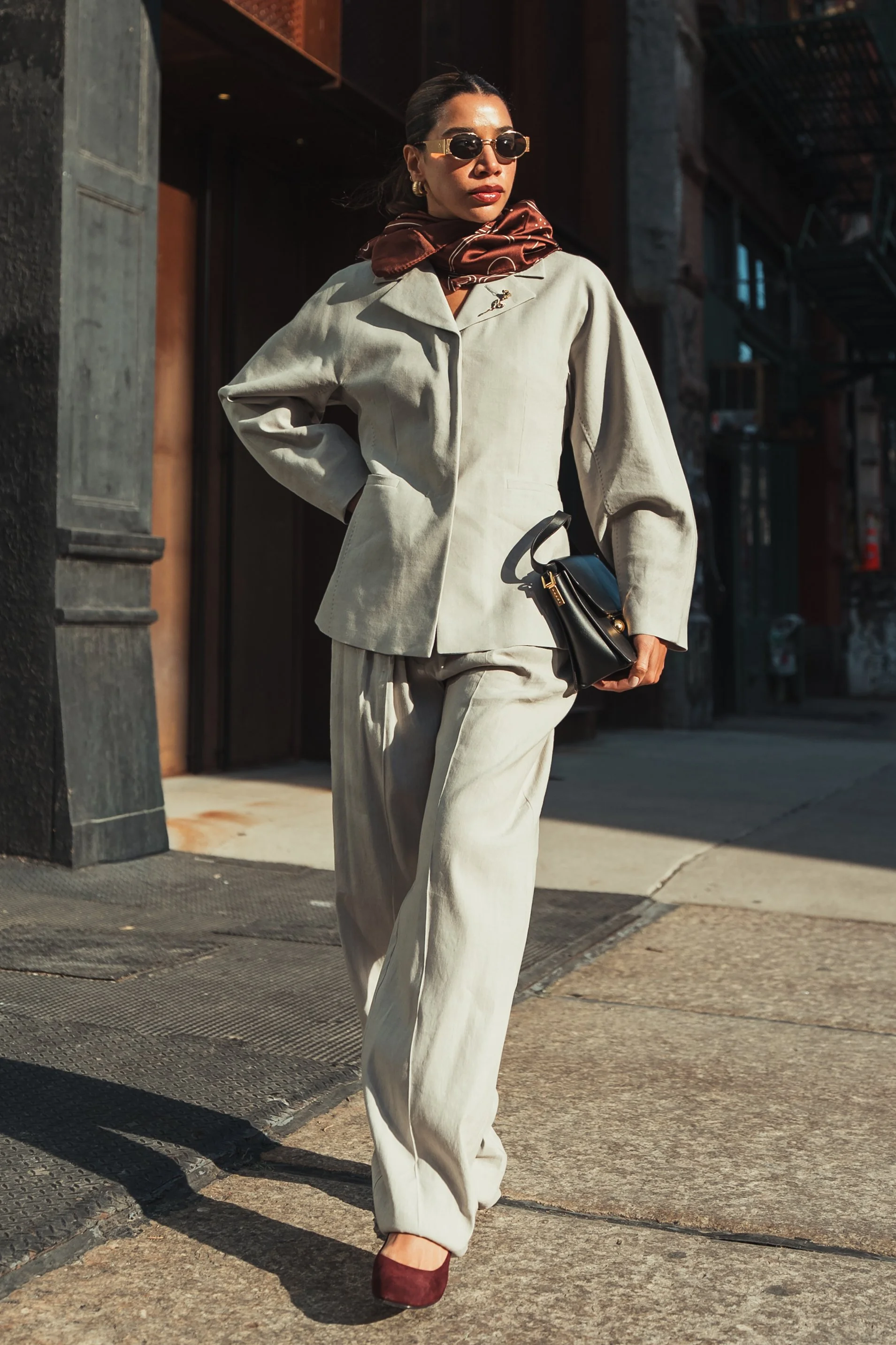 A stylish woman walking on a city sidewalk, dressed in a beige suit with a dark red scarf, sunglasses, and maroon shoes. She carries a black handbag and wears gold earrings.