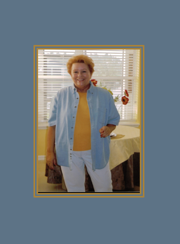Judy Goodman smiling indoors, standing in front of a window with blinds, wearing a yellow top, light blue button-up shirt, and white pants, with a table and floral arrangement in the background.