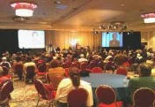 Judy Goodman teaching at a large conference room filled with people seated at tables, watching a presentation on a large screen at the front.
