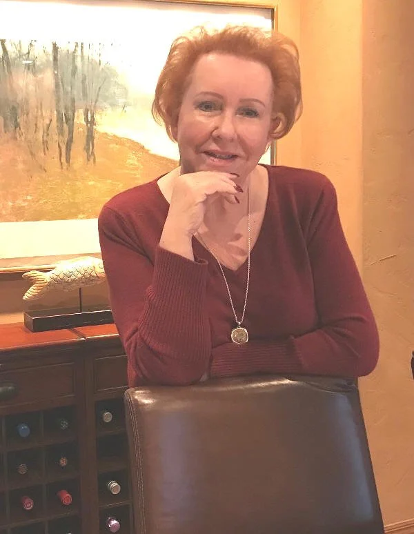 Judy Goodman wearing a maroon sweater and a pendant necklace, seated at a table in front of a painting of a tree-lined landscape.
