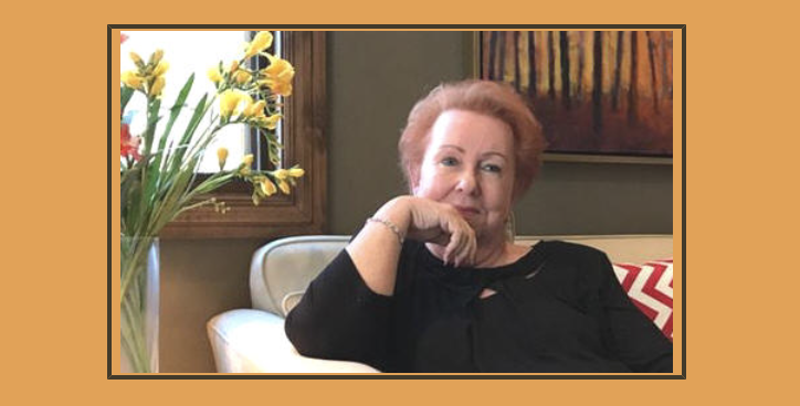 Judy Goodman with short red hair sitting on a white couch in a living room, with a bouquet of yellow and red flowers on a table beside her and a painting on the wall behind her.