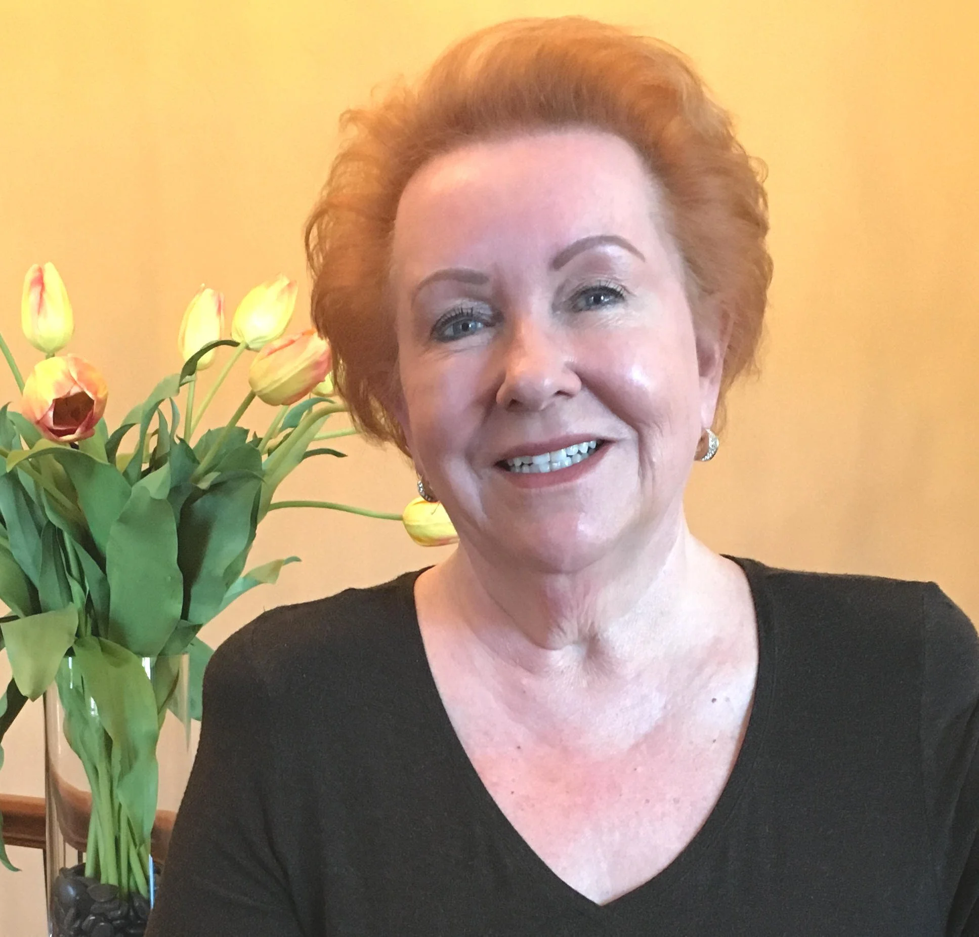 Judy Goodman with short, curly red hair posing in front of a bunch of yellow tulips with pink tips in a vase.