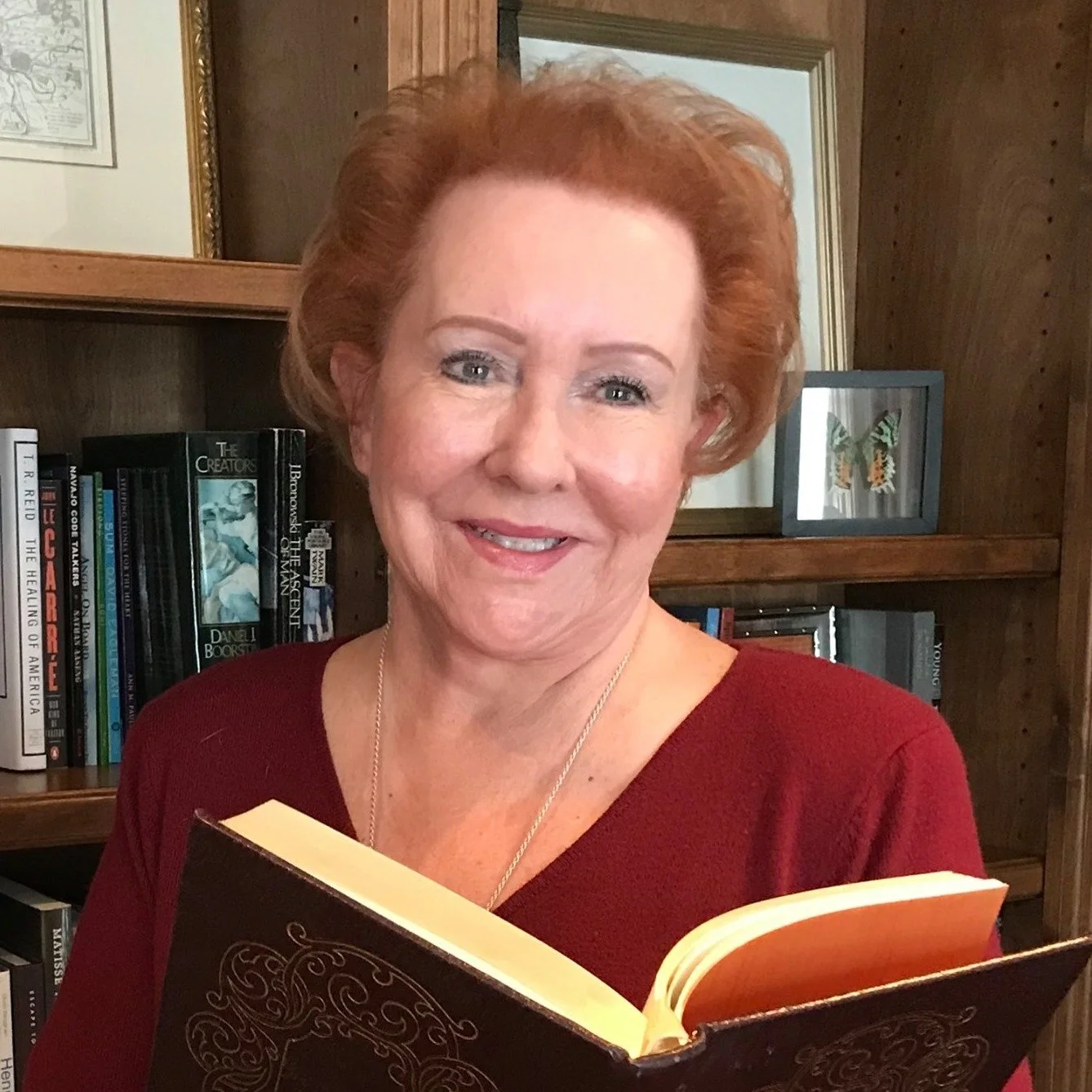 Judy Goodman, has short red hair smiling, holding an open book in a room with wooden bookshelves filled with books and framed pictures in the background.