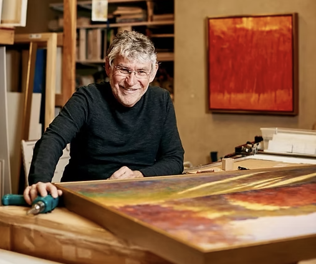 Artist Ken Elliott smiling while working on a large abstract painting in a home studio filled with art supplies and paintings.