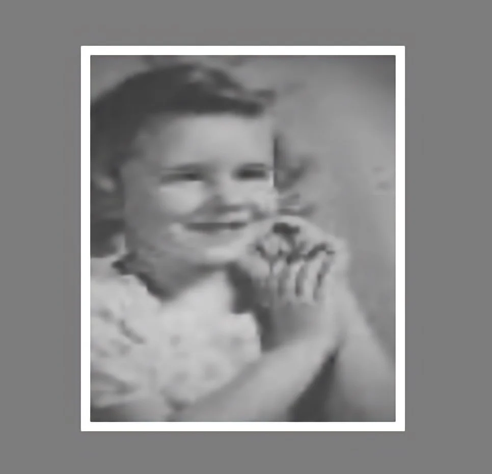 A black and white photo of Judy Goodman as a young girl smiling and resting her chin on her hand.