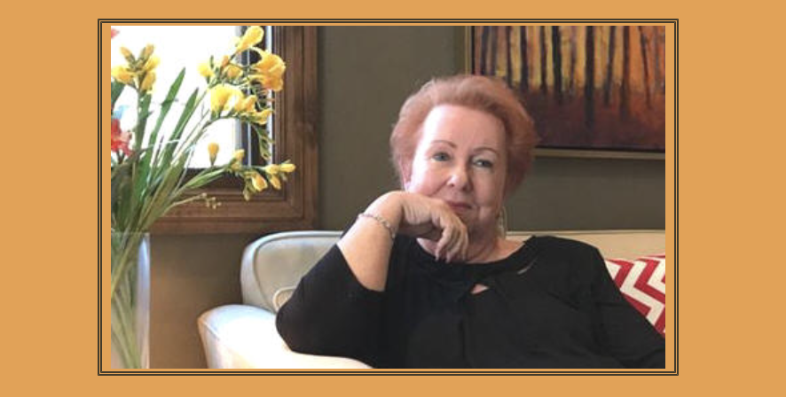 Judy Goodman sitting on a beige chair in a living room, with a vase of yellow flowers on a table nearby and a framed painting on the wall behind her.