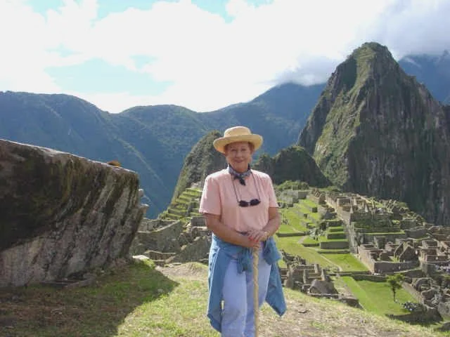 Judy Goodman standing in front of Machu Picchu ruins in the mountains, wearing a sun hat, pink shirt, and blue jeans, holding a walking stick.