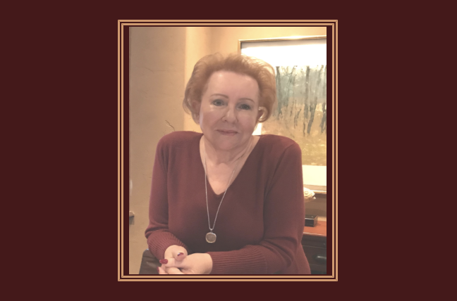 Judy Goodman with short, curly red hair sitting indoors, wearing a maroon sweater and a necklace, in front of a window with trees outside.