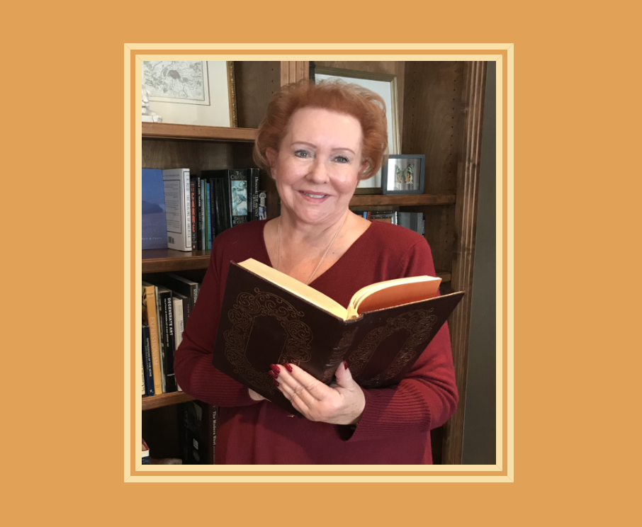 Judy Goodman holding an open book, standing in front of a wooden bookshelf with various books and framed pictures.