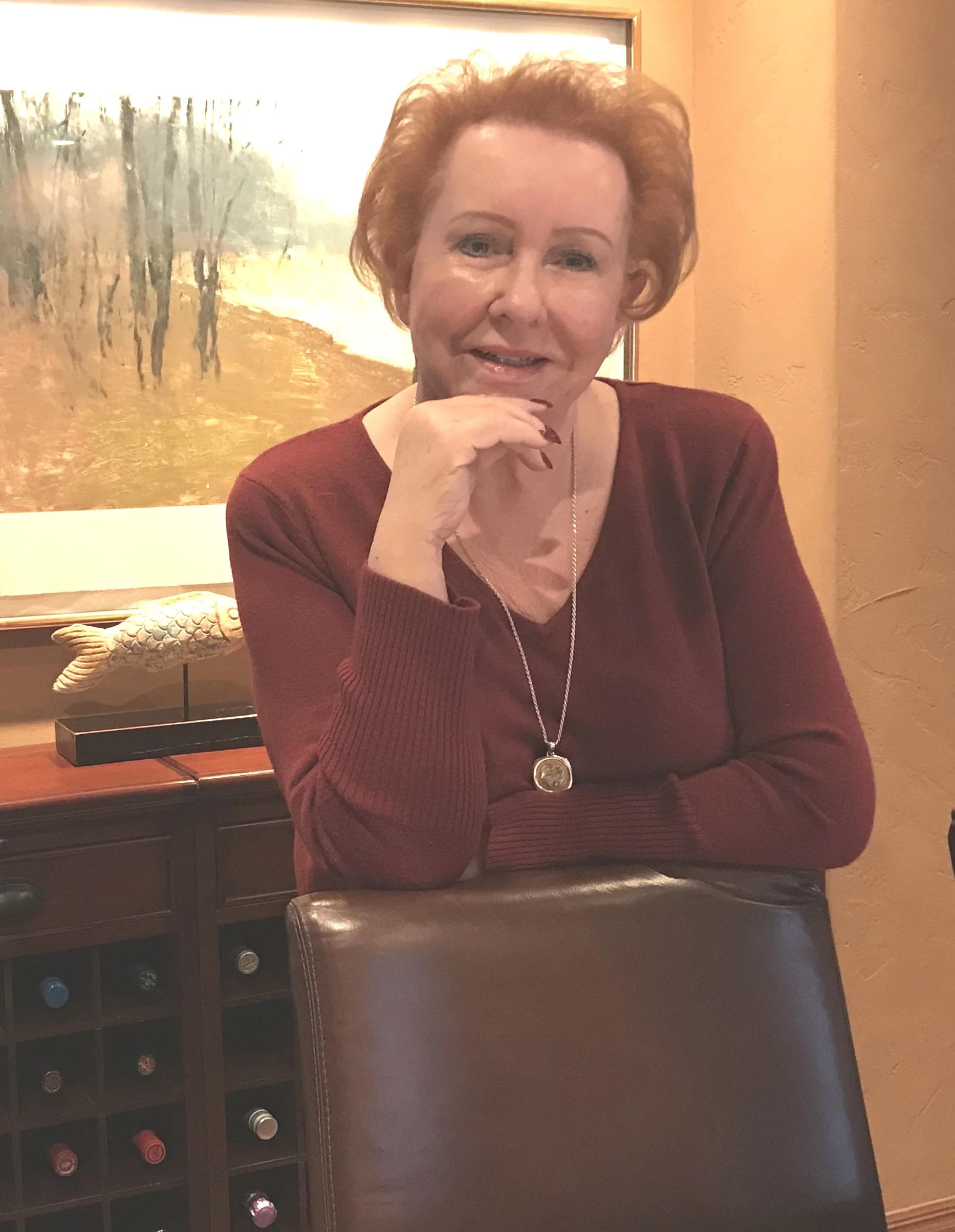 Judy Goodman wearing a maroon sweater and a necklace, sitting at a table in a room with a painting of trees and a wine rack in the background.
