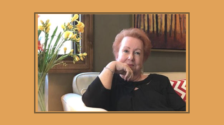 Judy Goodman sitting on a sofa near a window with yellow flowers in a vase, and a colorful painting on the wall behind her.