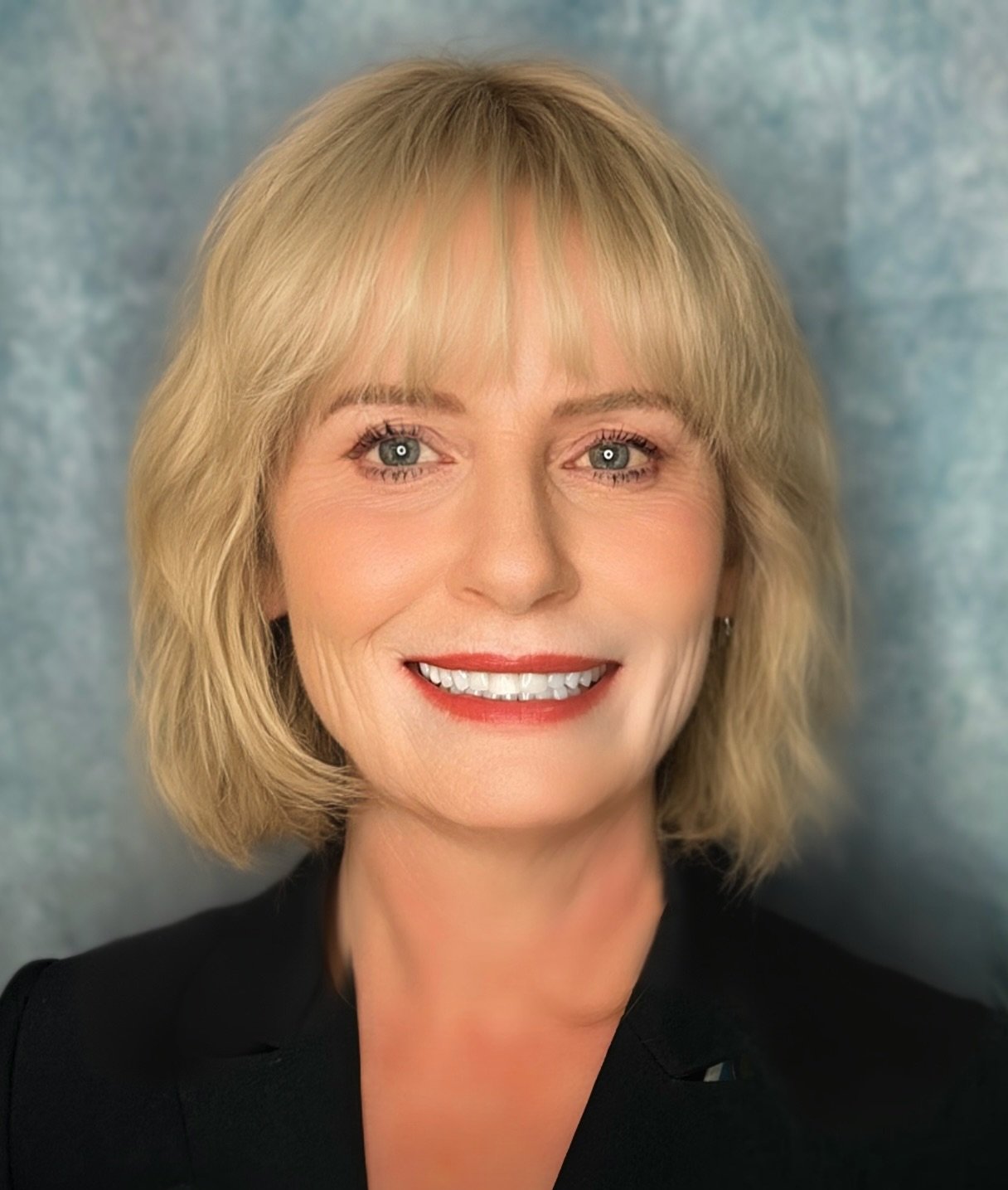 A woman with blonde hair and blue eyes smiling, wearing a black top against a blue-gray background.