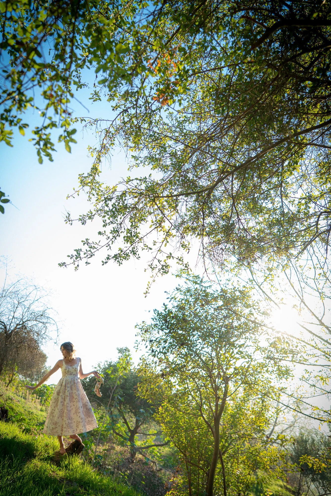 A woman in a white floral dress standing on grassy terrain surrounded by trees with green leaves, sunlight shining through the branches, creating a bright, outdoor setting.