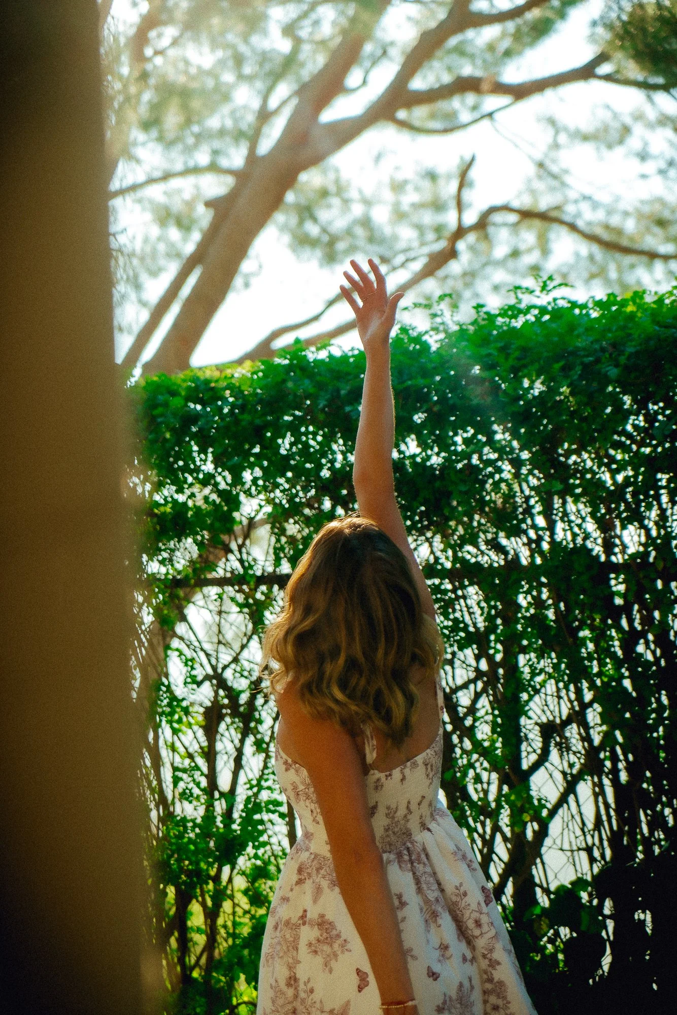 A woman with wavy brown hair wearing a floral dress raising her arm in front of green foliage and trees.