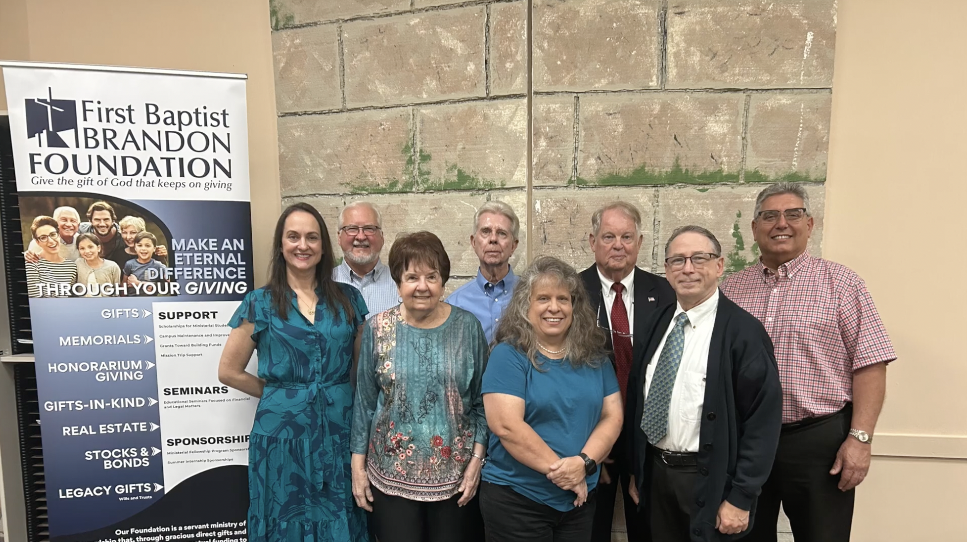 Group of nine people standing in front of a wall and a First Baptist Brandon Foundation poster at a fundraising or community event