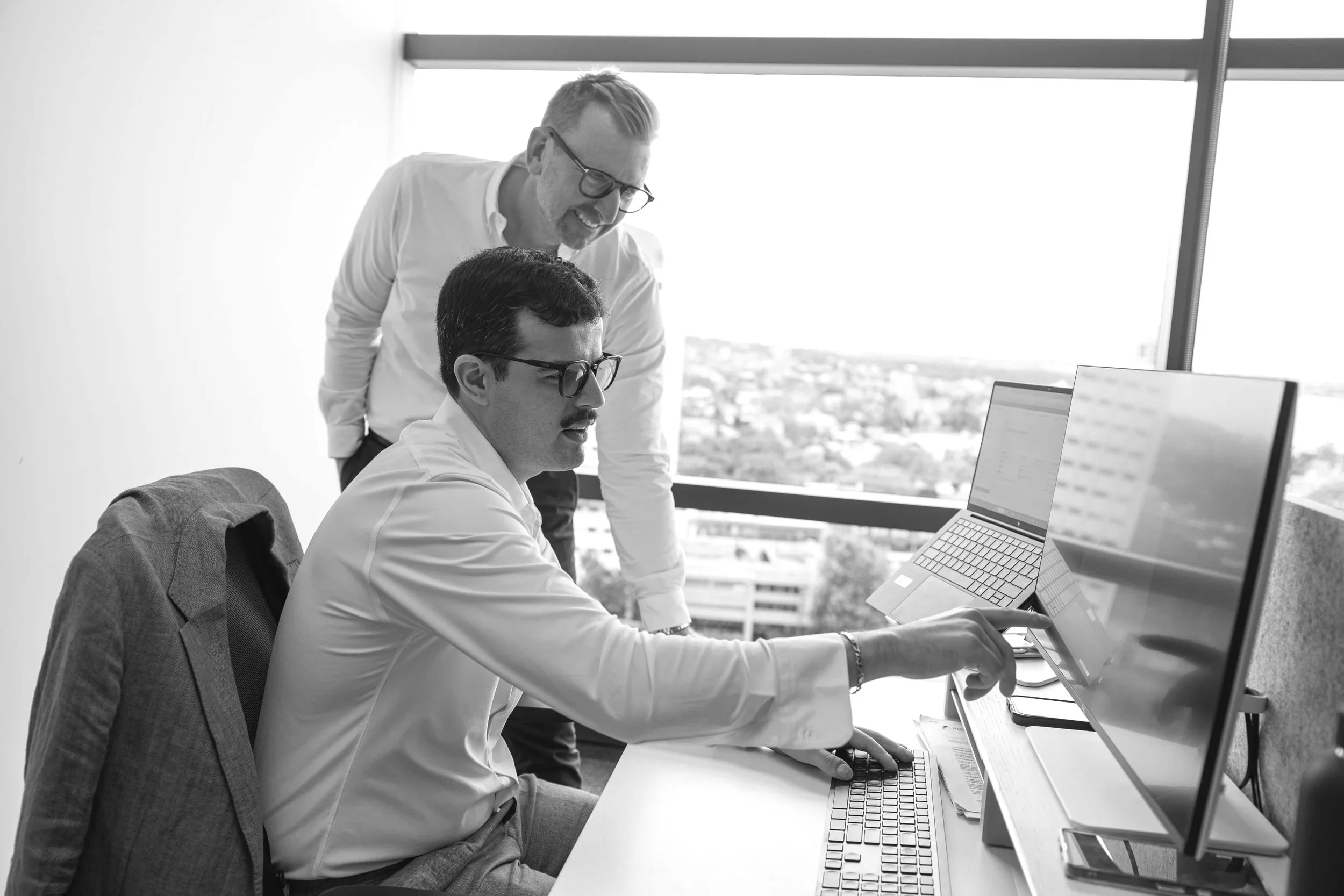 Two men working together at a computer in an office with large windows, one is sitting and pointing at the screen while the other stands behind him smiling.