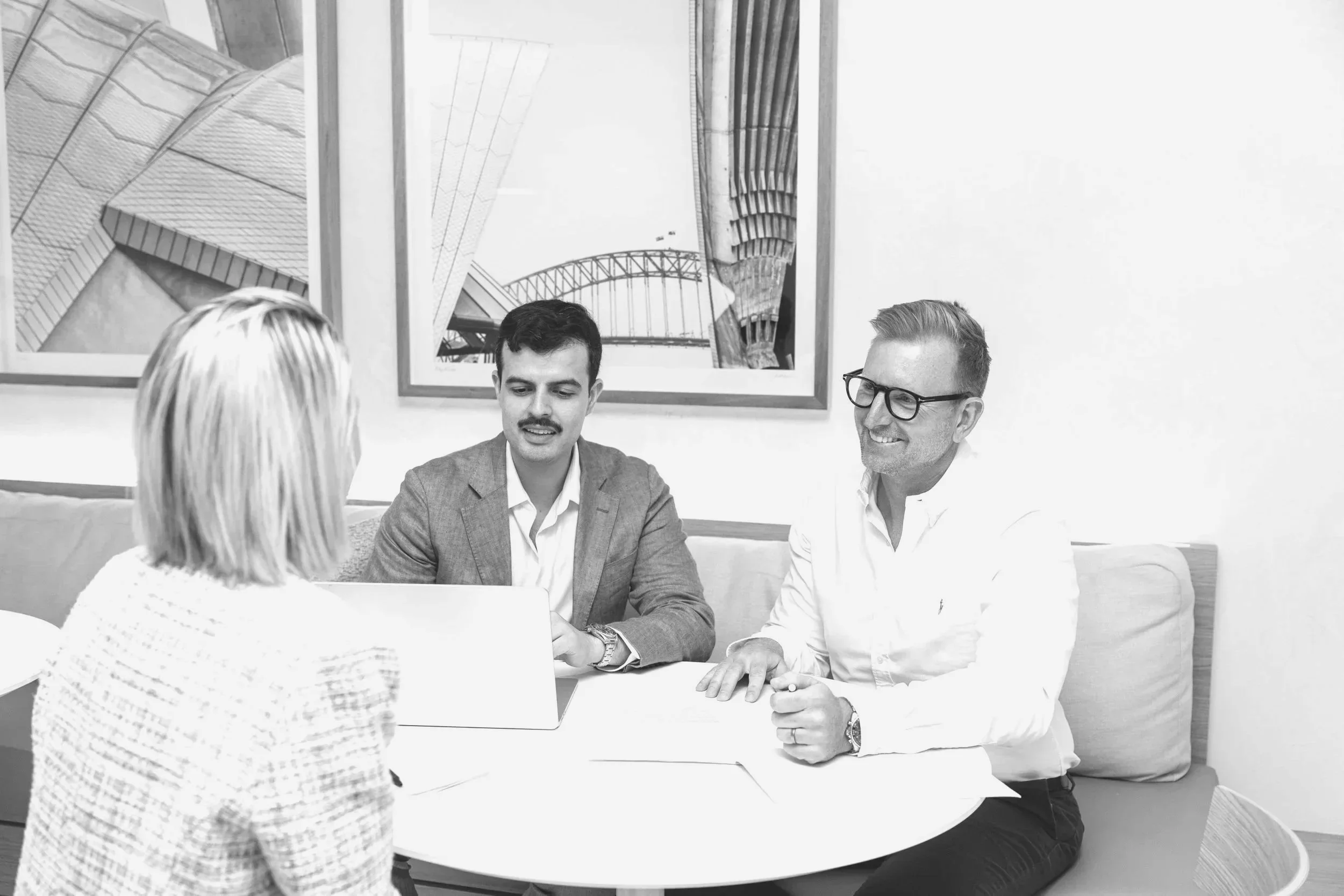 A group of three people in a business meeting, sitting around a round table with laptops, in an office setting with framed architectural photos on the wall.