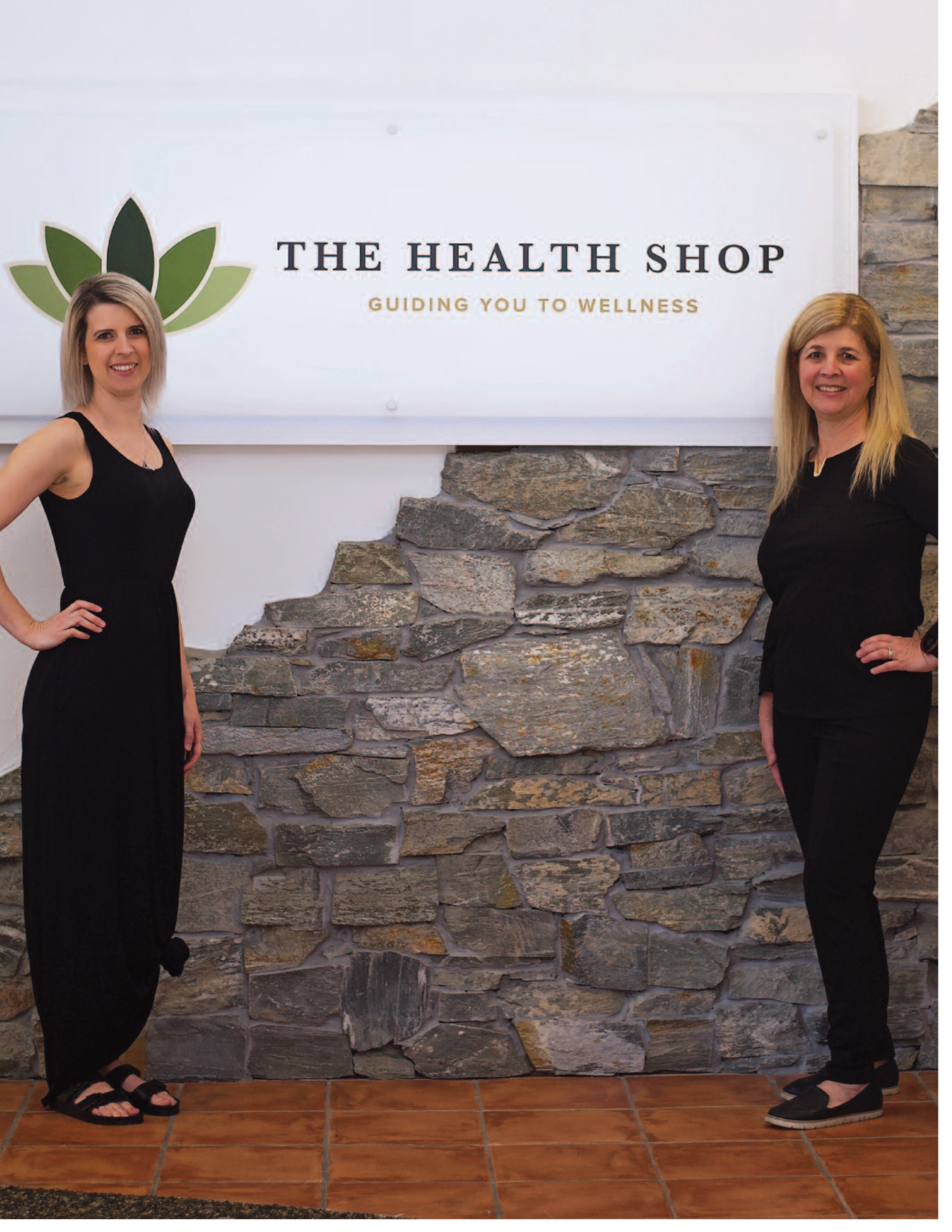 Two women standing on either side of a sign for The Health Shop, with a stone wall background and tile flooring.