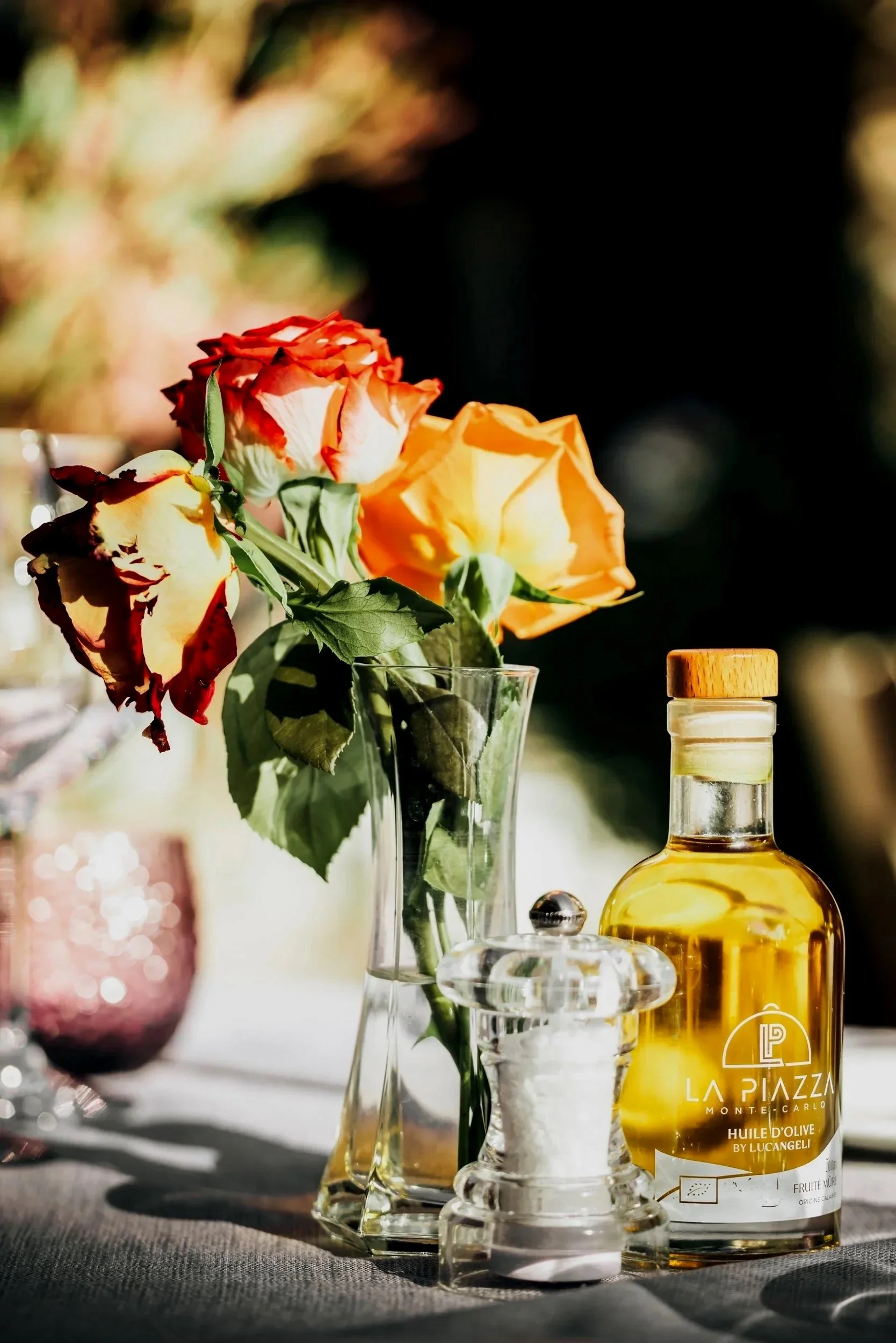 Close-up of a bouquet of roses in a glass vase, with a bottle of olive oil in the foreground, on a table with sunlight streaming in.