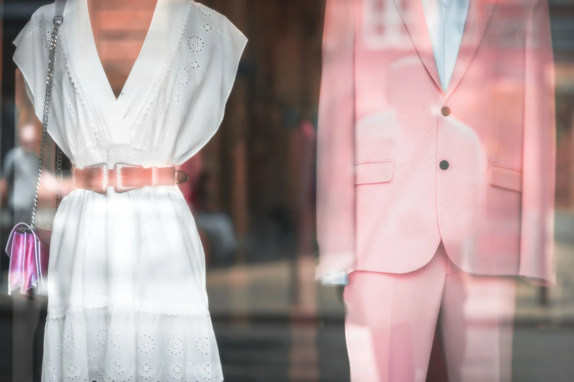 Store window display featuring a woman wearing a white dress with eyelet embroidery and a pink belt, and a man wearing a pink suit with a white shirt underneath.