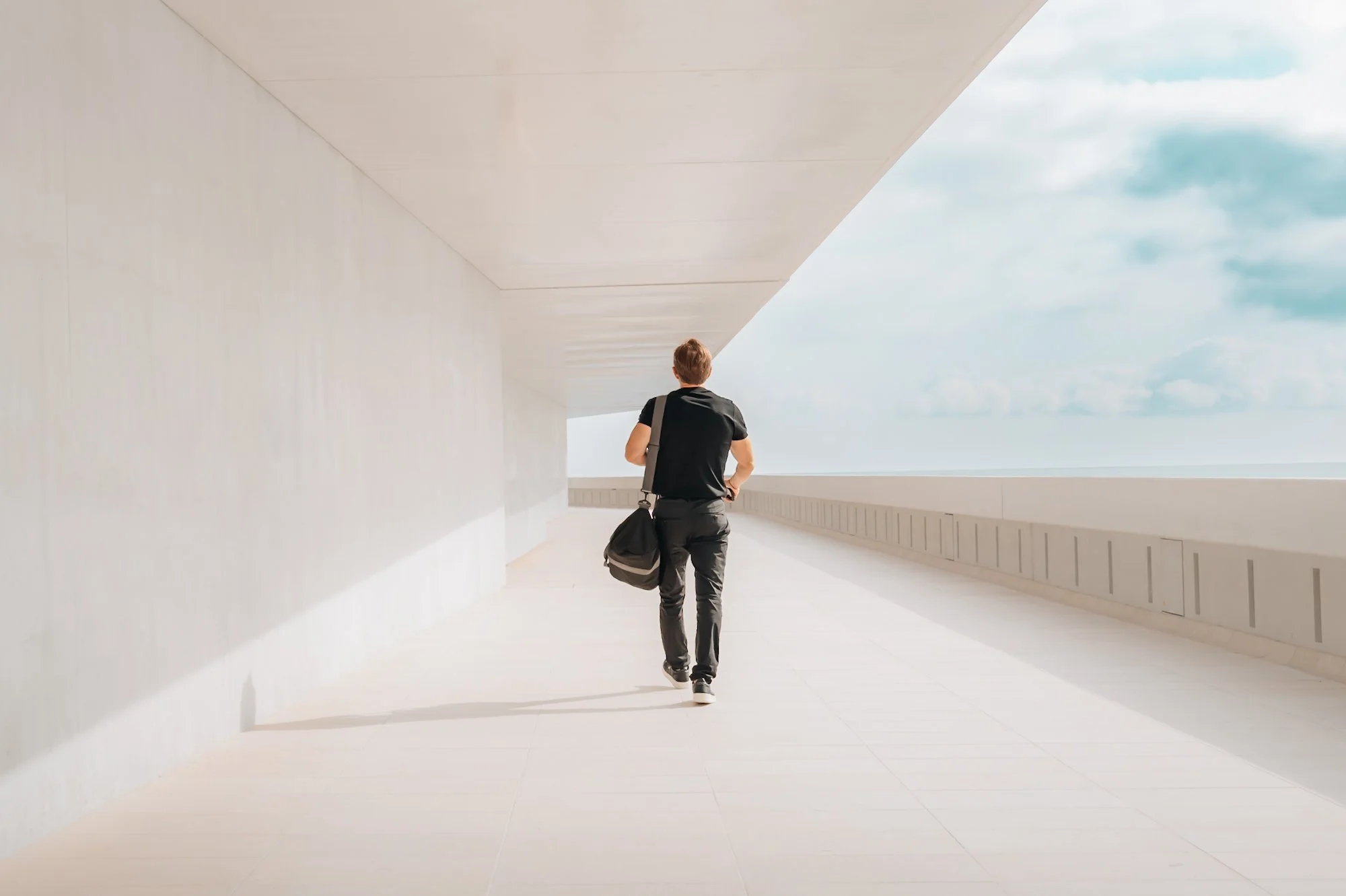 A person walking in a modern, minimalistic white corridor with a curved ceiling, holding a black bag, with an open view of the sky and clouds outside.