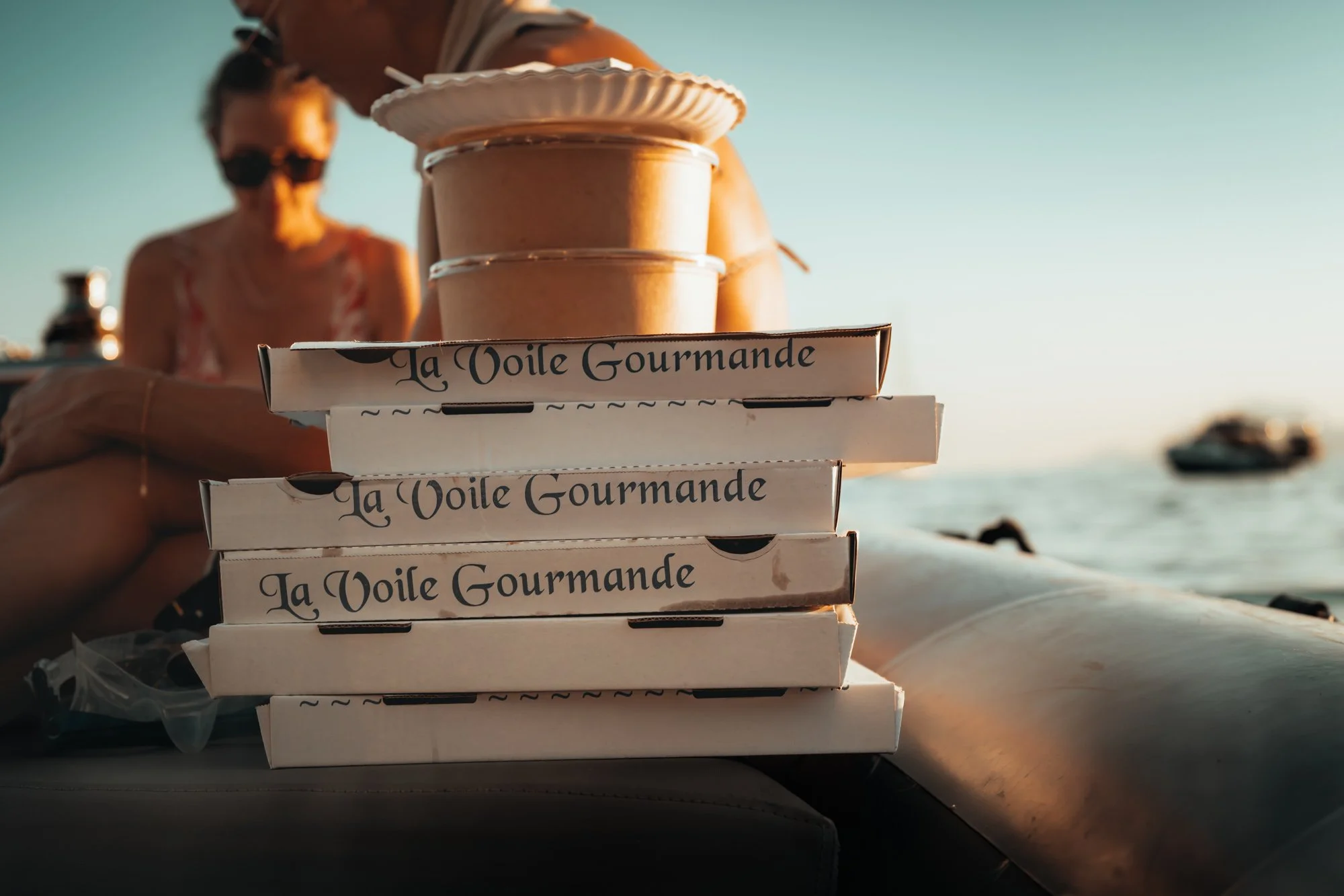 The image shows a stack of white pizza boxes with elegant print that says "La Voile Gourmande". There are three small containers of food stacked on top of the pizza boxes. A woman wearing sunglasses is in the background, on the beach, with the ocean 