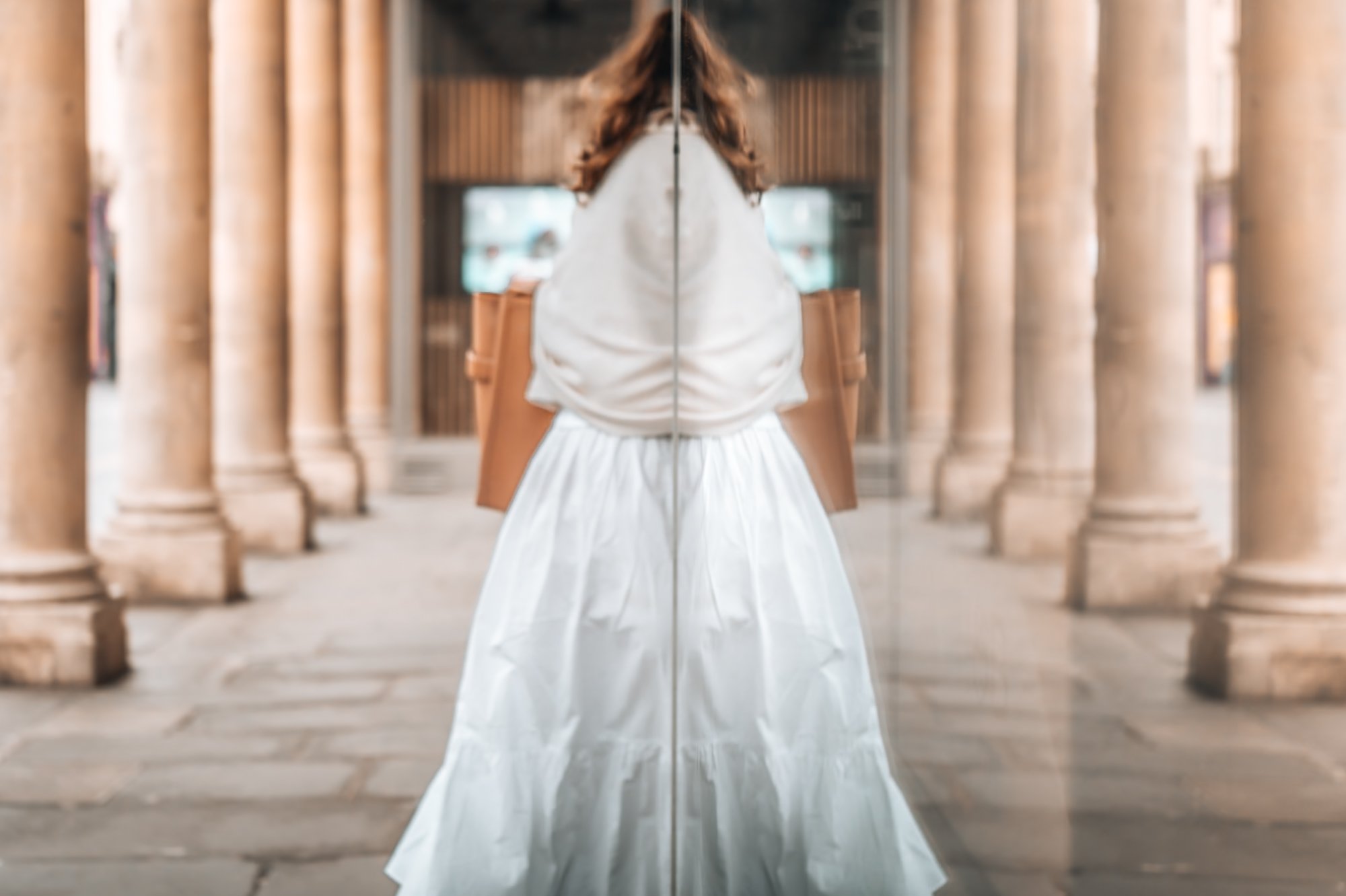 A woman with auburn hair in a white dress stands behind a mirror, reflecting her back. She is in an architecturally grand corridor with tall columns on either side.
