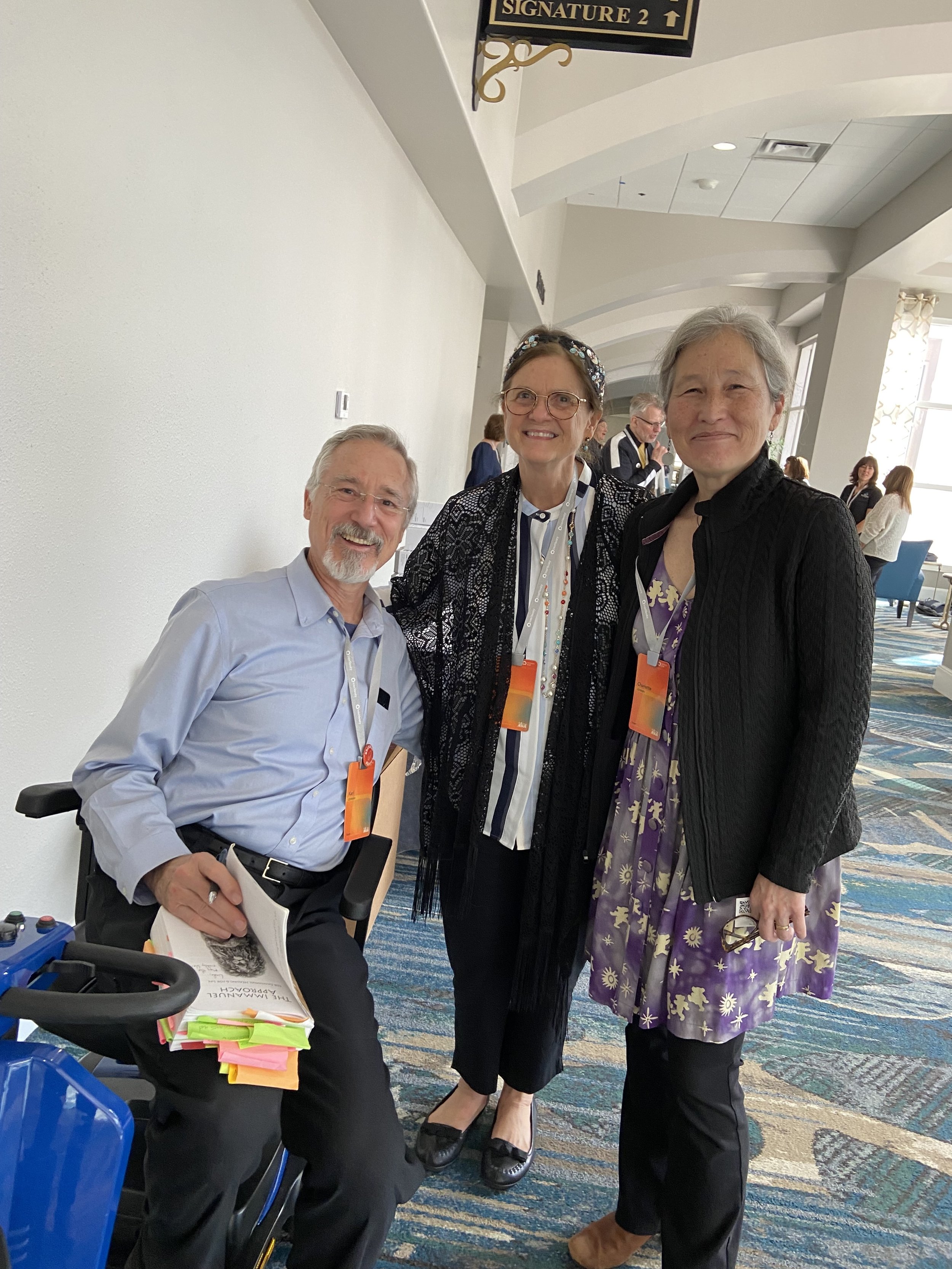 Three people smiling and posing for a photo at a conference or event, with name tags around their necks. The setting appears to be a brightly lit, modern indoor space with a patterned carpet and white walls.