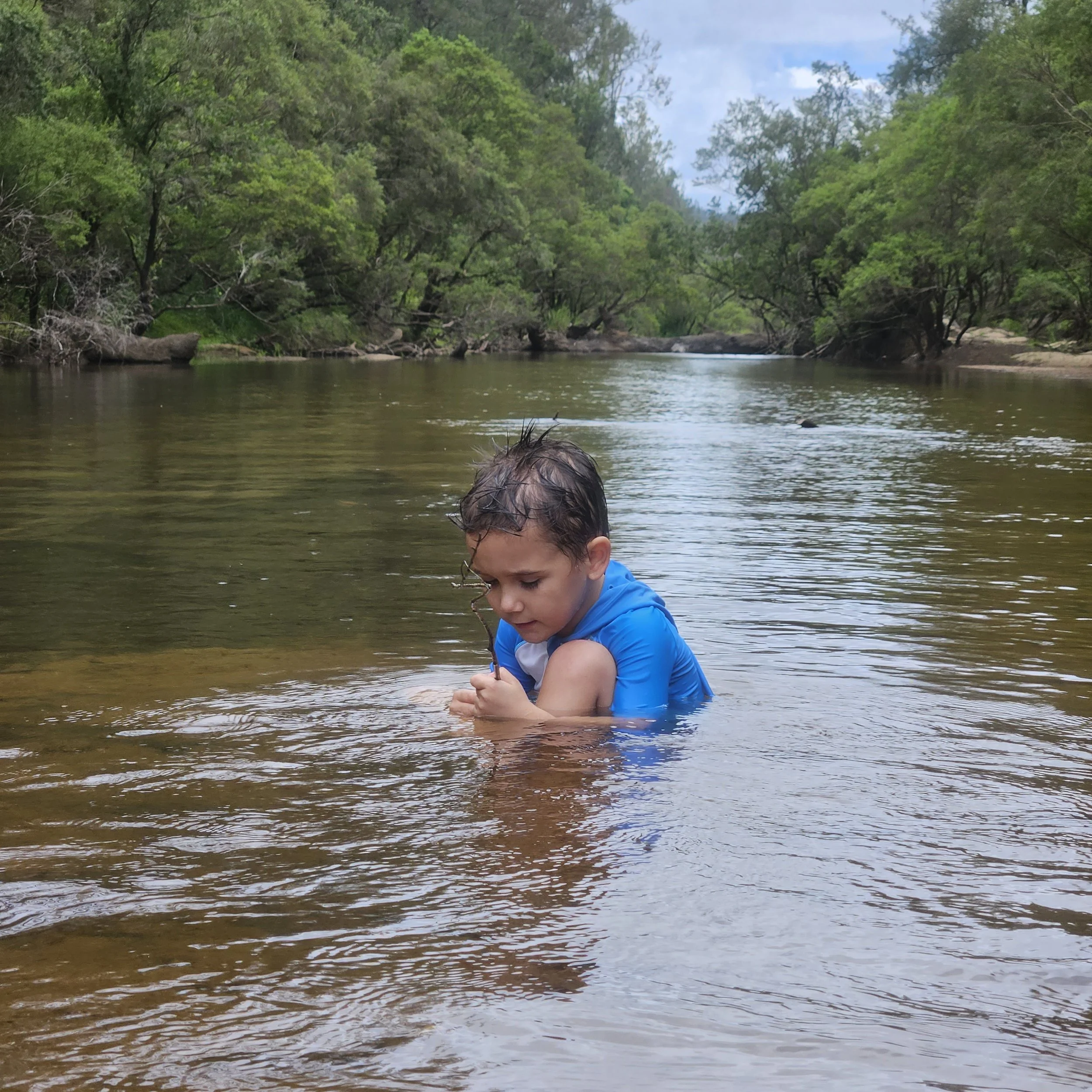 Young boy with wet hair and a blue shirt playing in a shallow river surrounded by green trees.