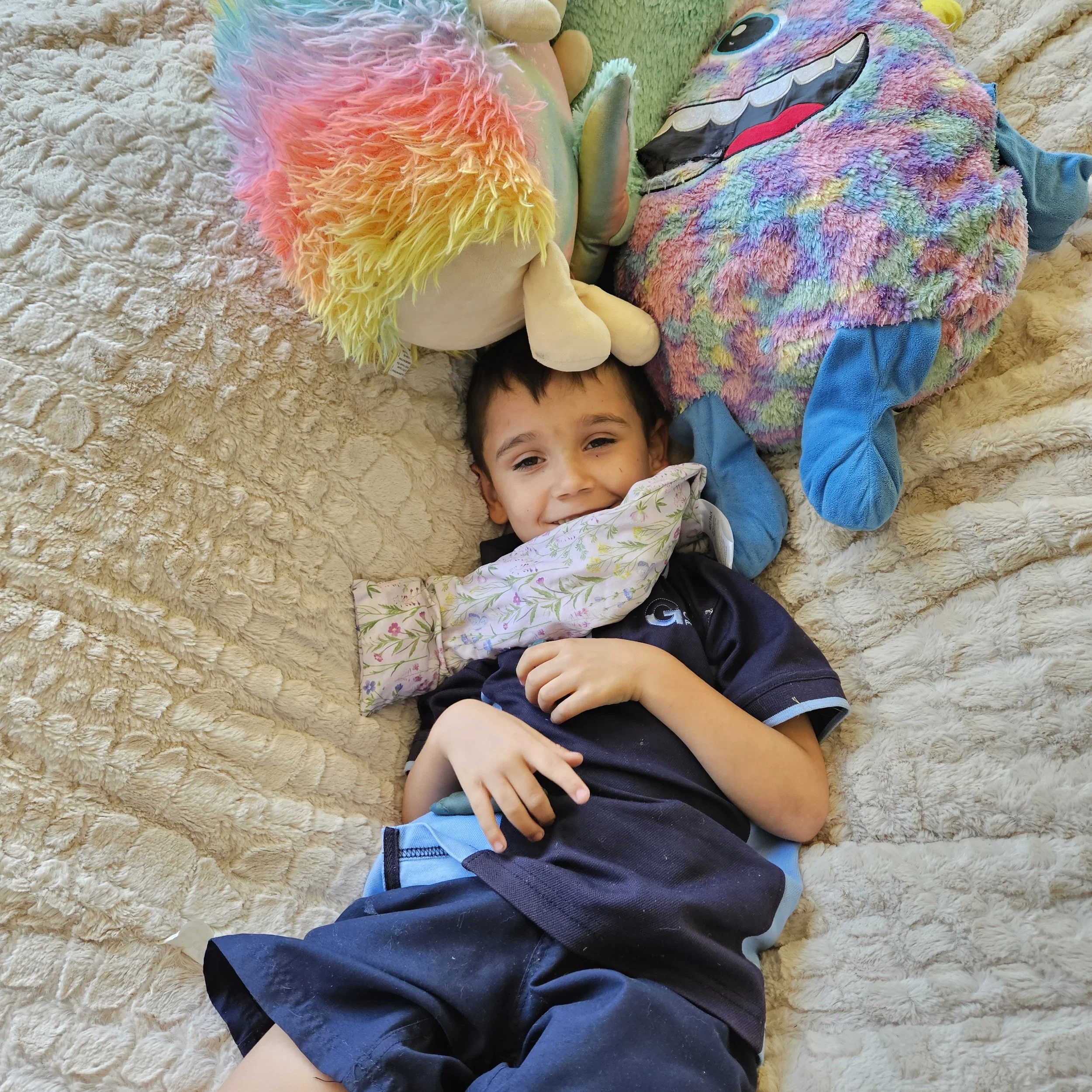 A young boy lying on a beige textured blanket, smiling and surrounded by colorful stuffed animals, including a rainbow fluffy plush and a pastel-colored unicorn.