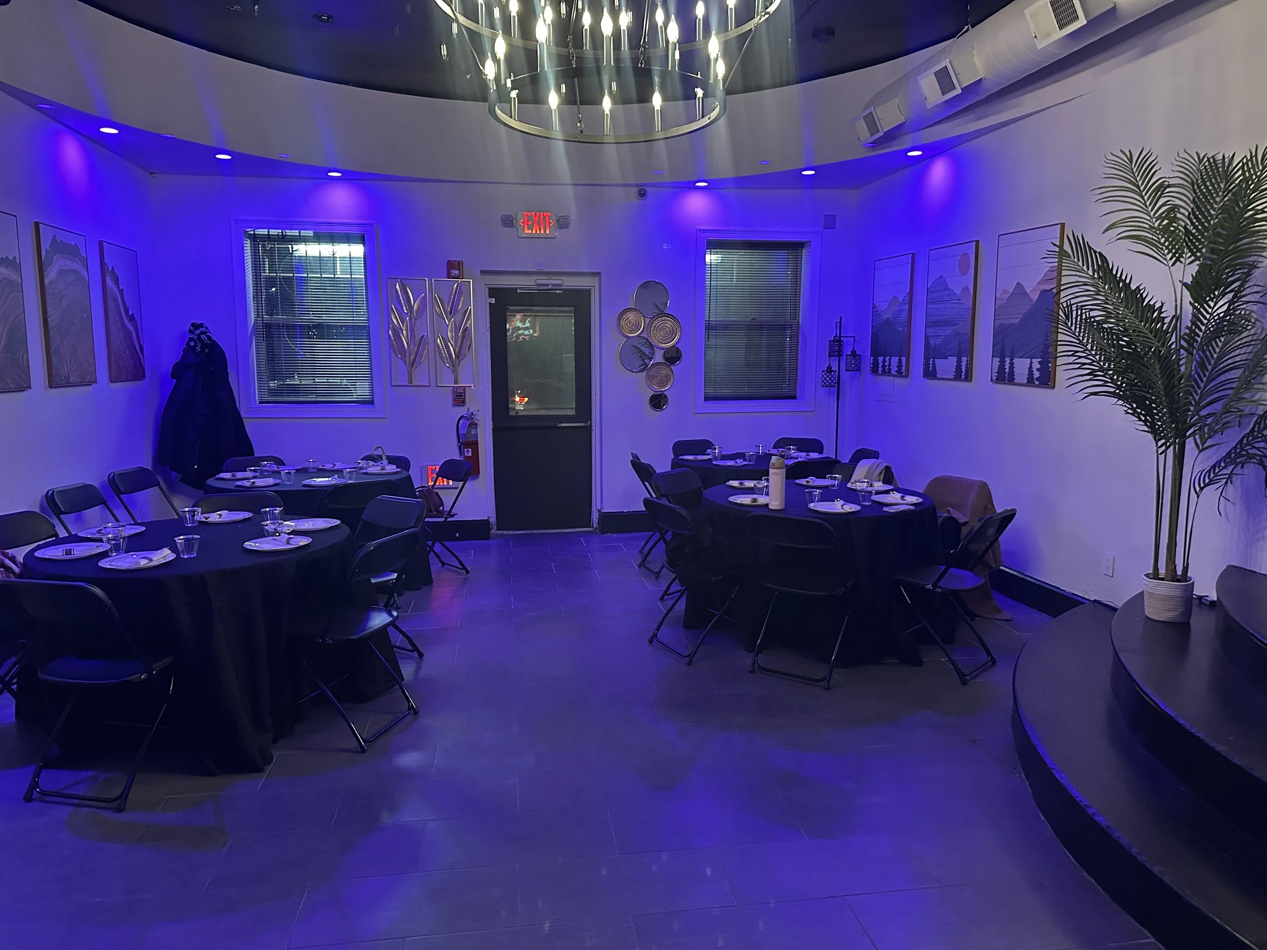 An empty indoor dining room with round tables covered in black tablecloths, set with plates, glasses, and utensils. The room has blue ambient lighting, several framed landscape artworks on the walls, and a large potted plant in the corner.