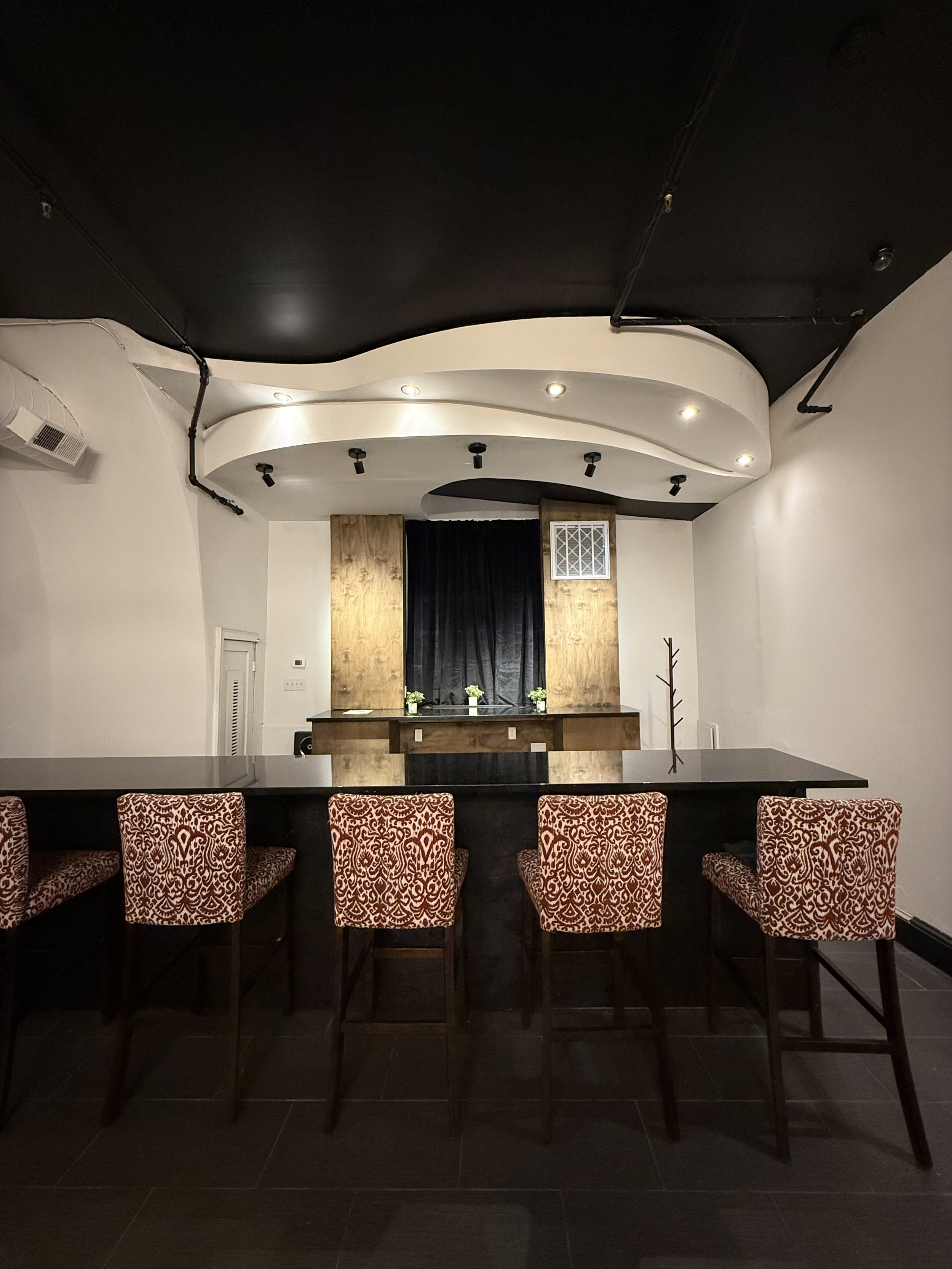 Interior of a bar or lounge with a black countertop, five patterned chairs, a small stage with black curtains, wood paneling, and minimal decoration.