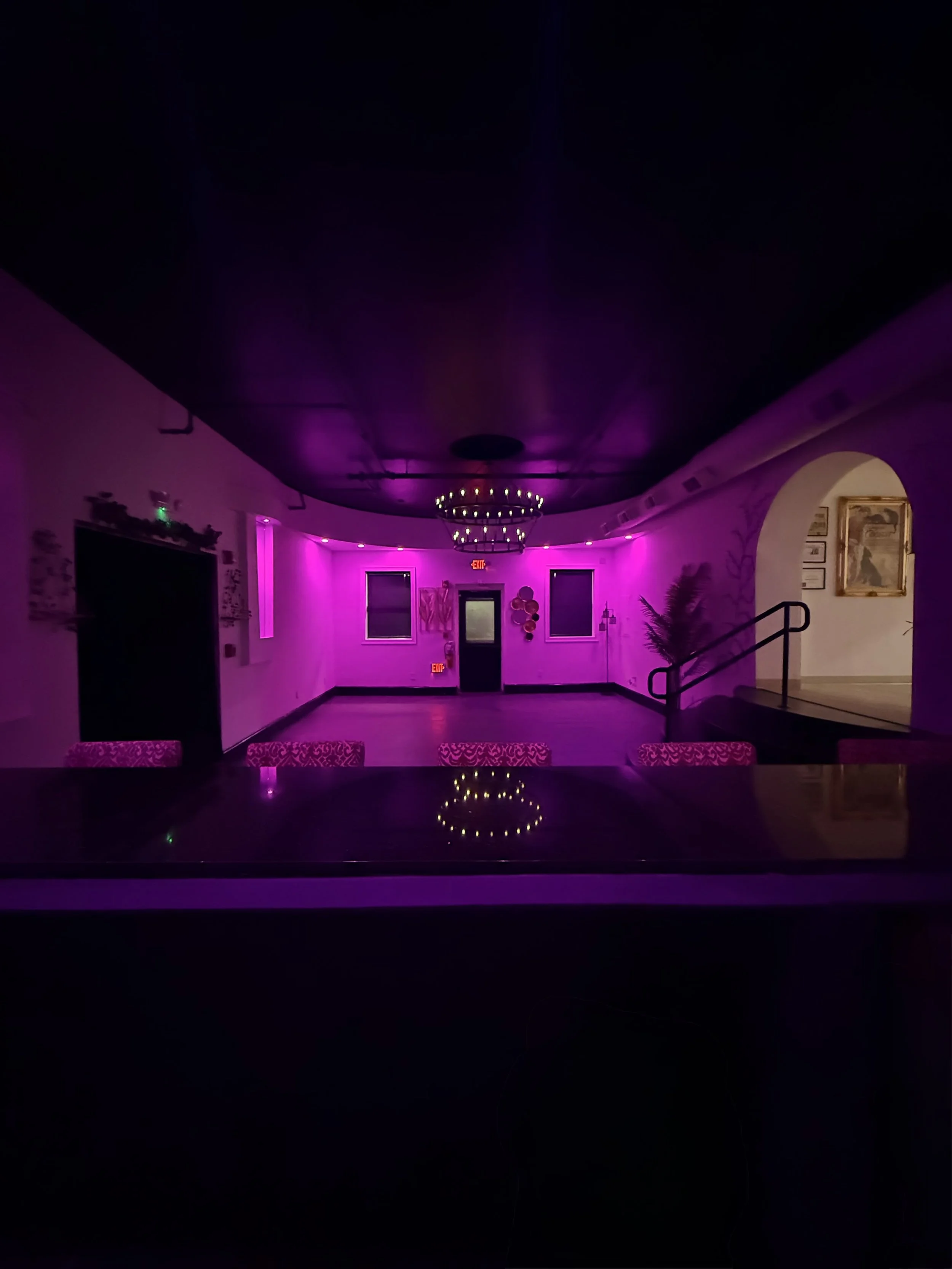 Empty banquet or event hall illuminated with pink and purple LED lights, decorated with wall art, plants, and chandeliers.