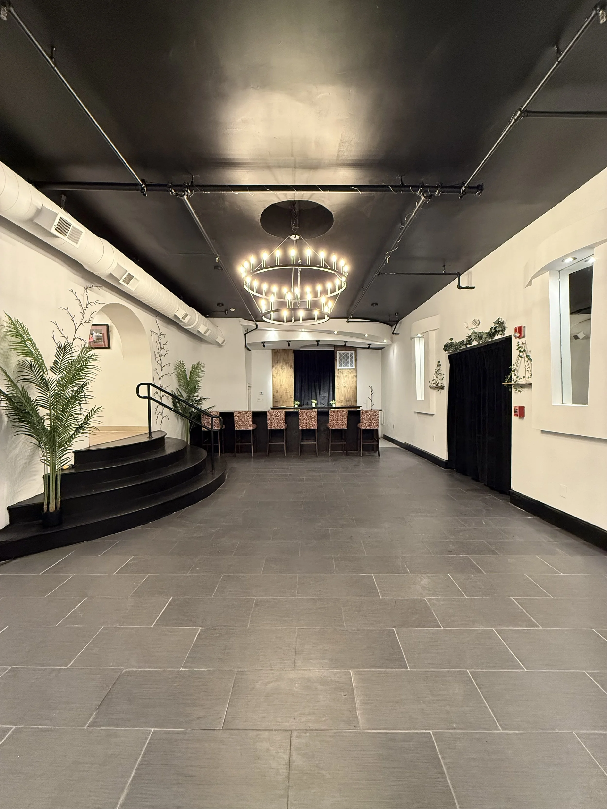 Empty event space with black ceiling, chandelier, plants, and a bar area with high chairs.