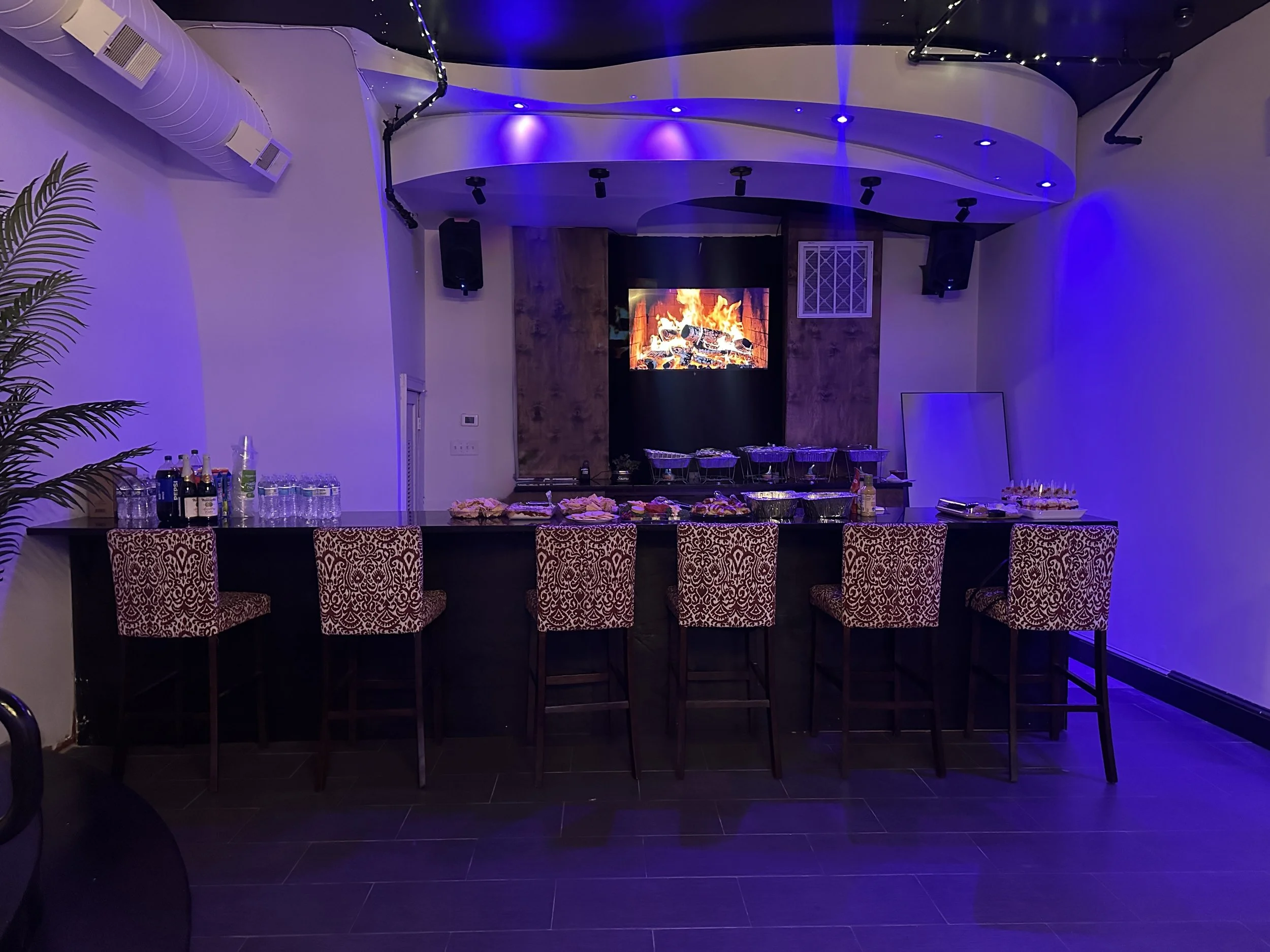 Party bar setup with bottles, glasses, and snacks in a room with purple lighting and a digital fireplace on a TV screen.