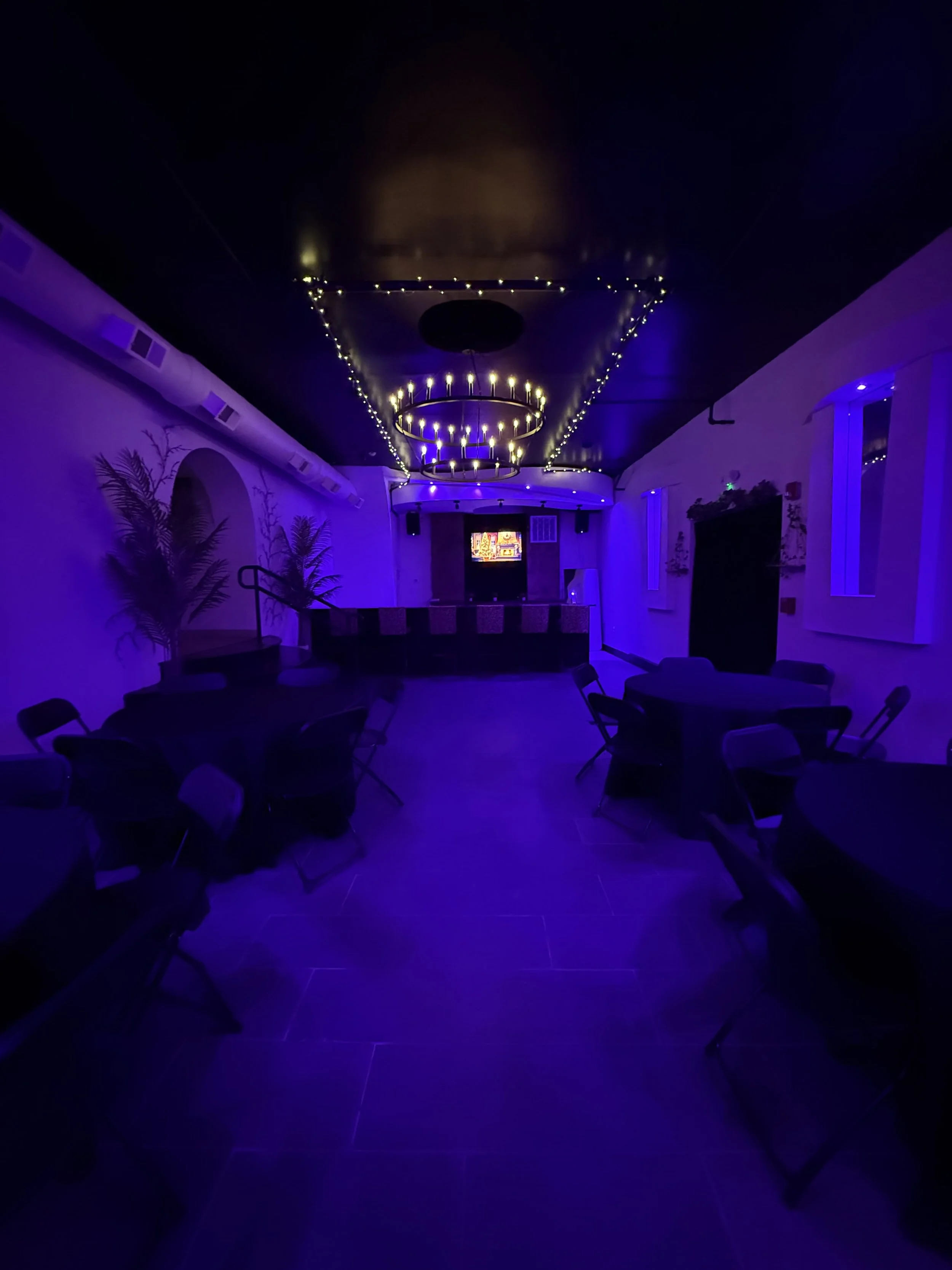 Dimly lit banquet hall with purple lighting, round tables with chairs, chandelier with candles, and decorative wall art