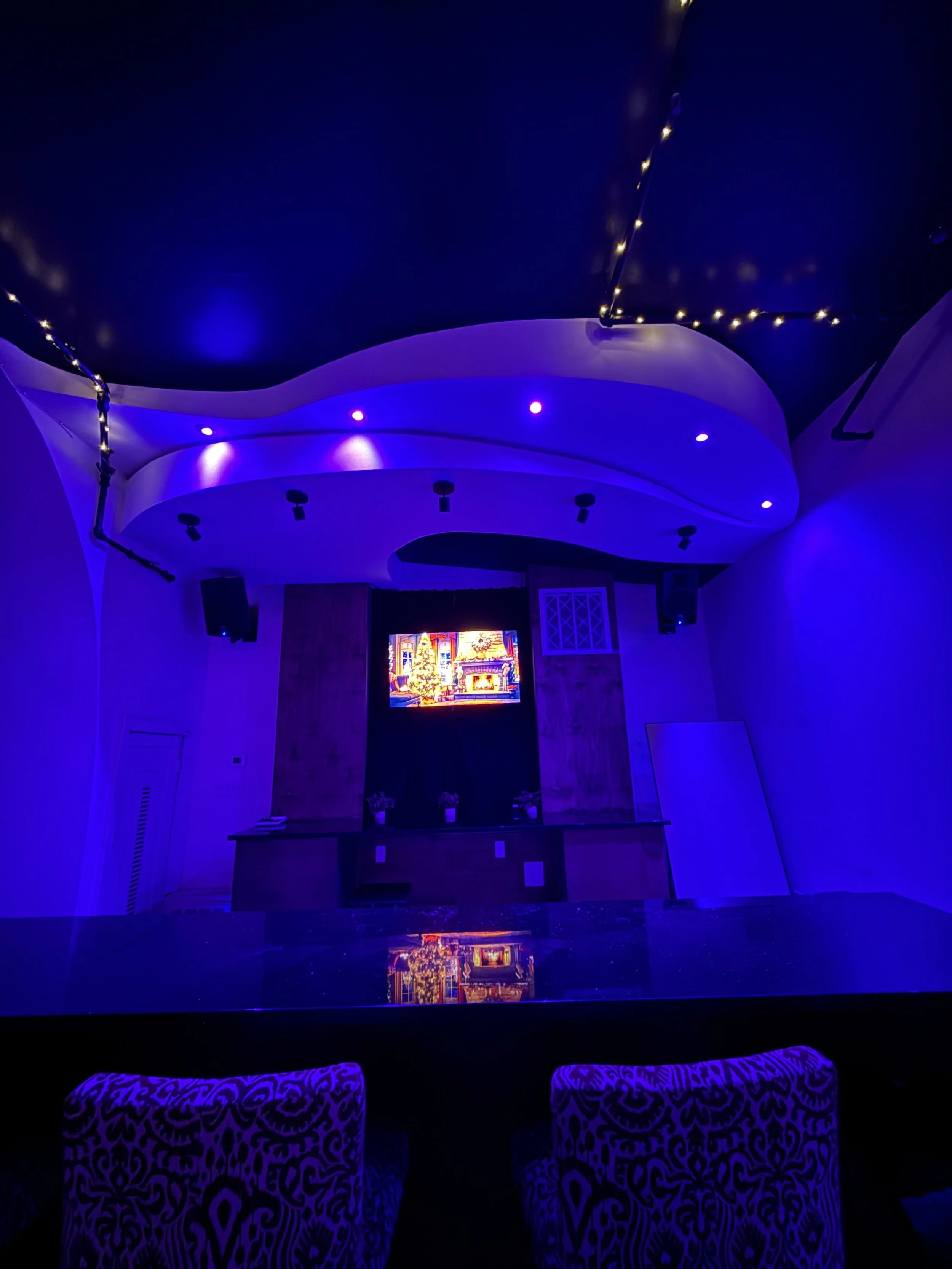 Indoor stage with Christmas tree and decorations, illuminated in blue lighting, featuring a television screen showing a festive scene, surrounded by chairs and string lights.