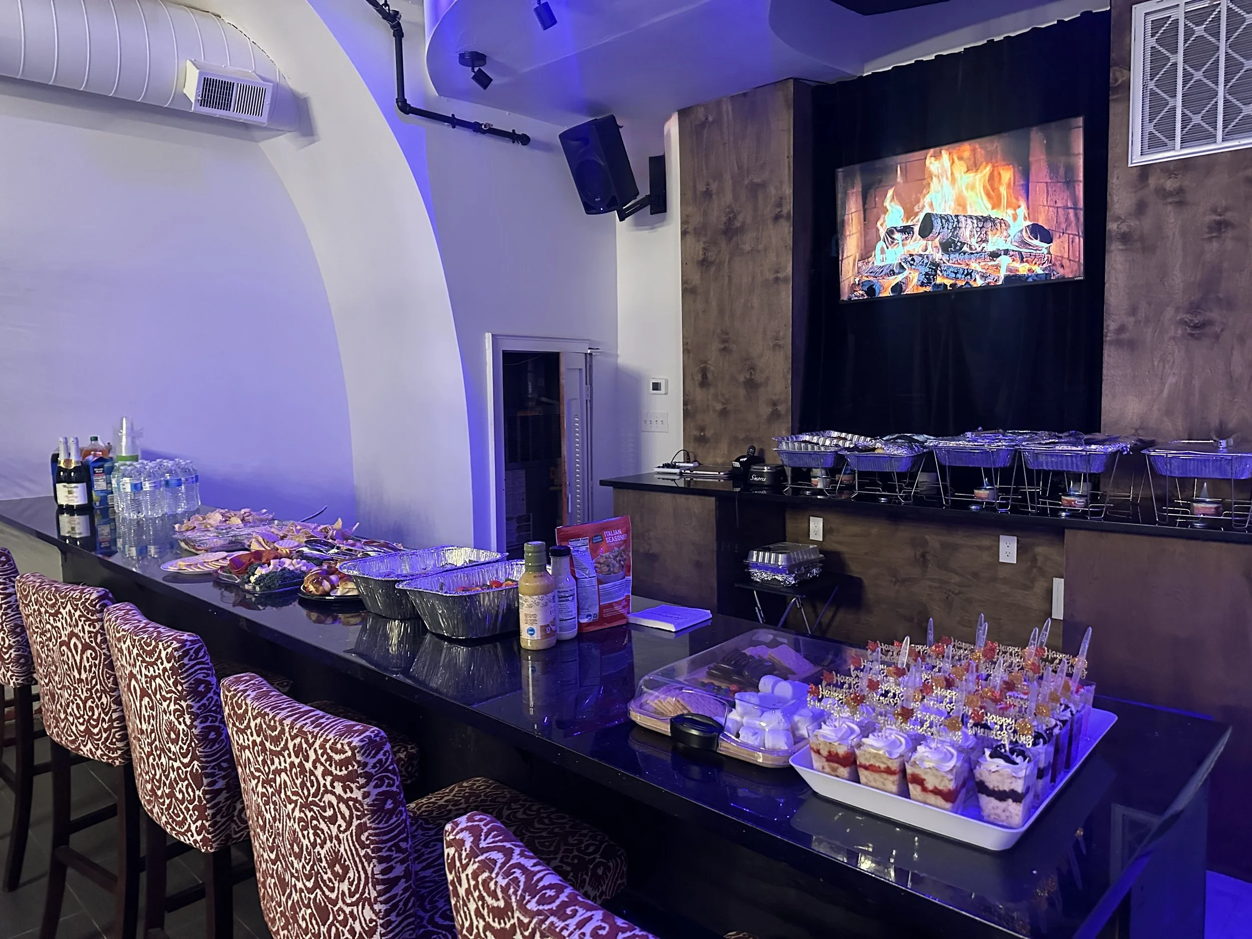 Party room setup with a long black table filled with food and drinks, pink patterned chairs, and a large flat-screen TV displaying a fireplace, creating a cozy atmosphere.