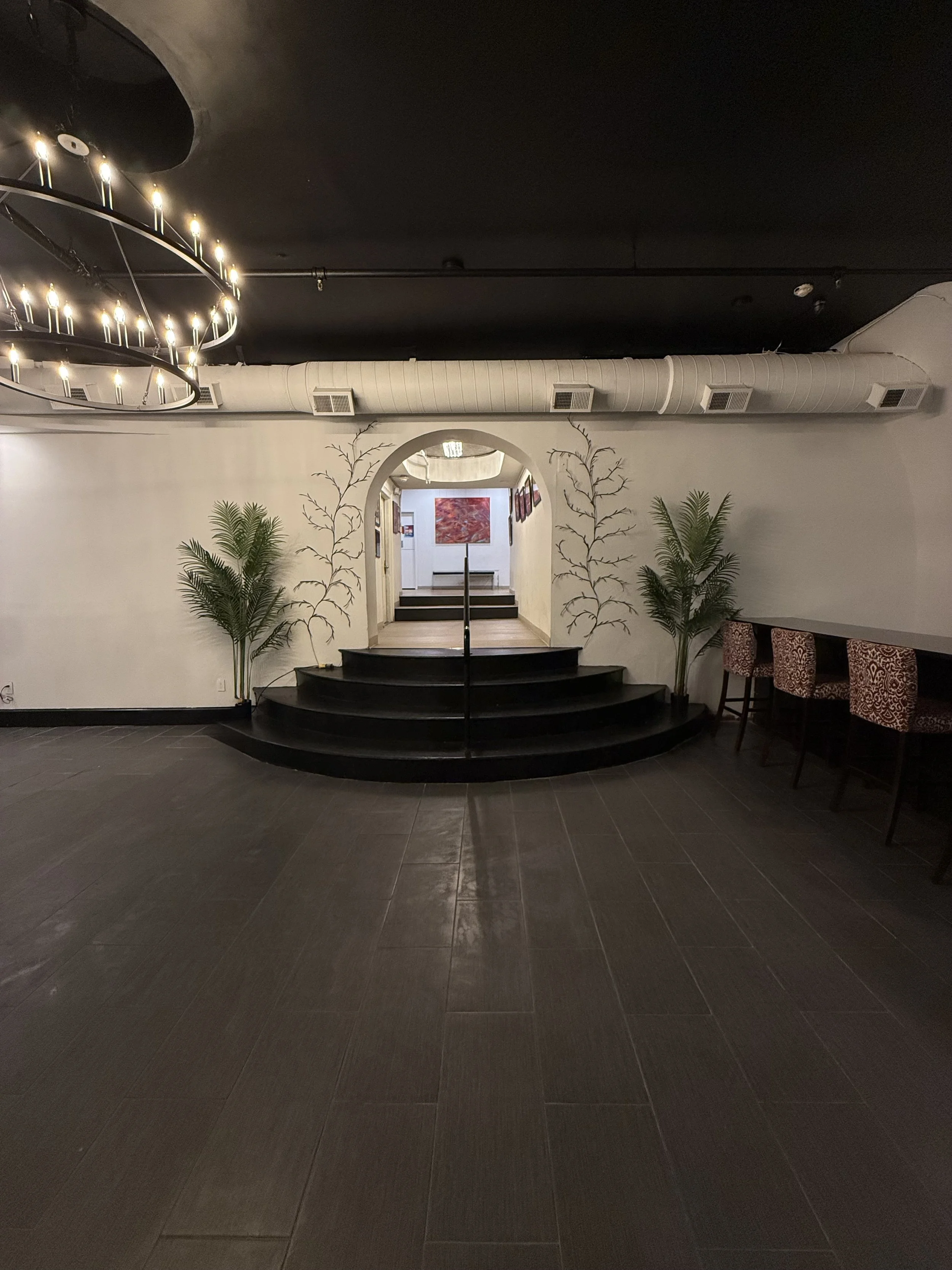 Interior view of an art gallery or restaurant with black tumbled tile flooring, white walls, a small staircase leading to another room, decorative plants, and wall art, with a dark ceiling and modern chandelier.