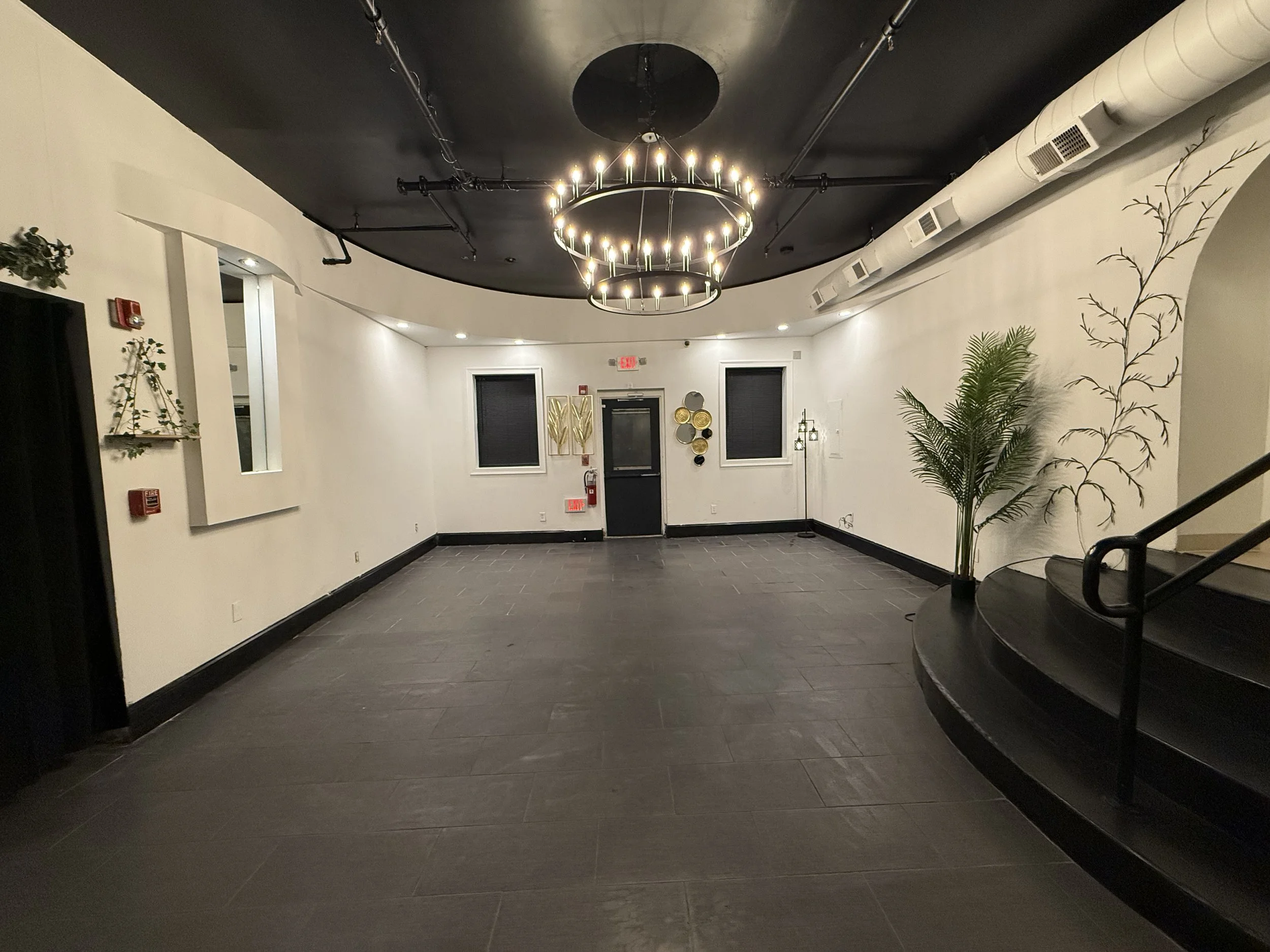 Empty modern event space with dark tile flooring, white walls, black ceiling with exposed ducts, and a chandelier. Decor includes wall art, plants, floor lamp, and windows with black blinds.