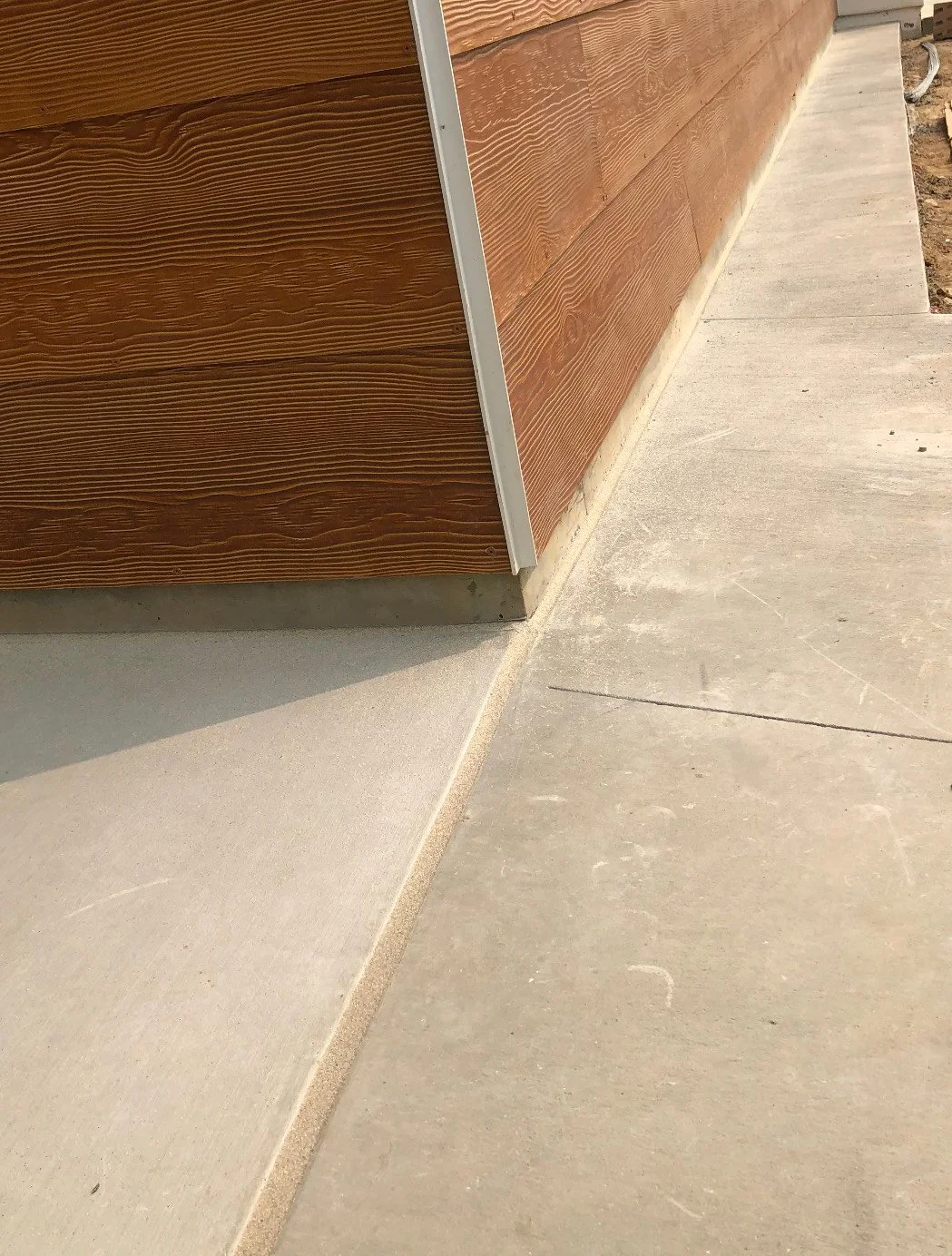 Close-up of a building's exterior corner, showing wooden siding, aluminum trim, and concrete sidewalk with expansion joint.