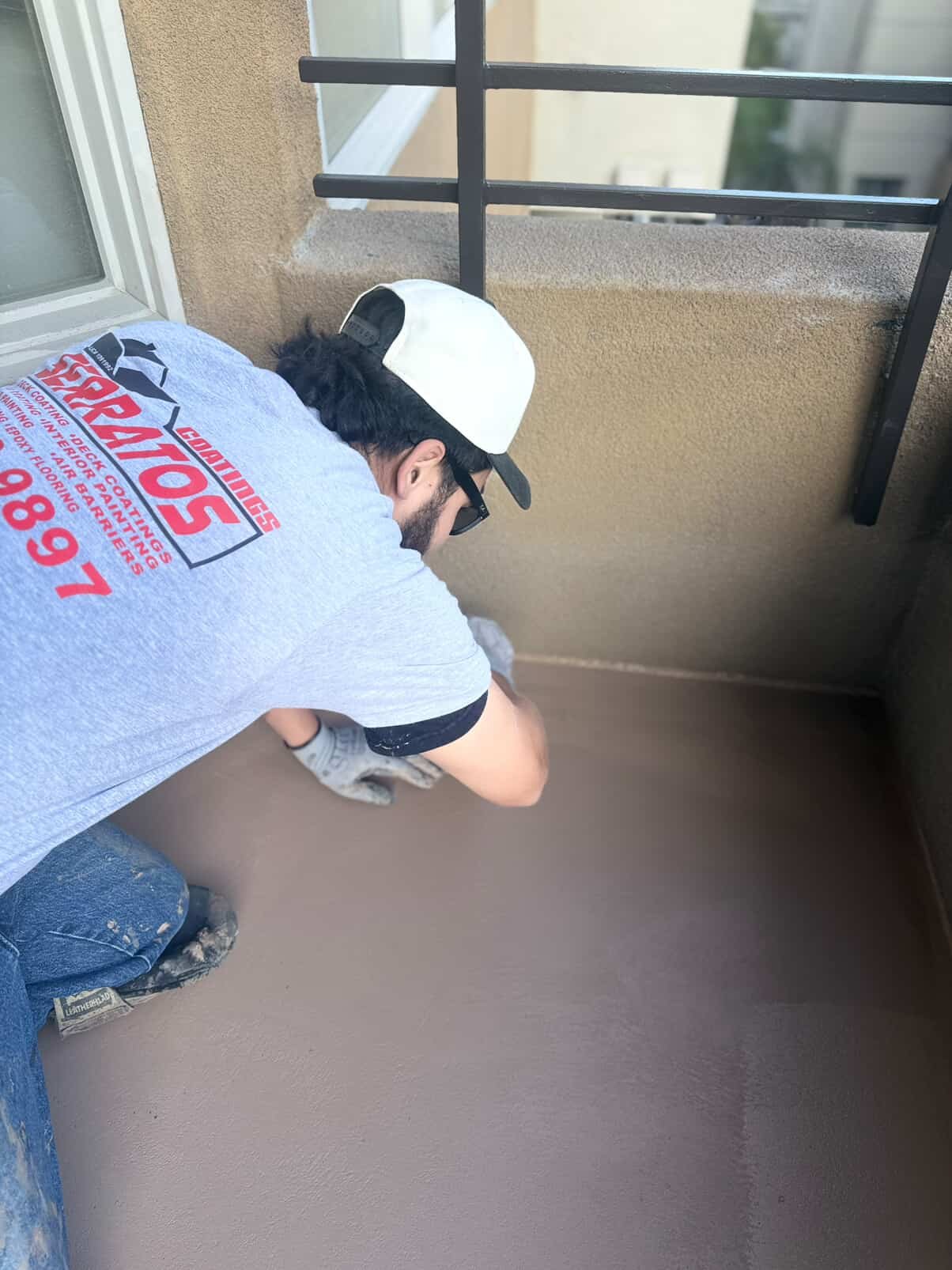 A man crouching on a balcony, working on a small dog. He is wearing a gray T-shirt, a white cap backwards, and glasses. The balcony has a beige wall, a window, and a black metal railing.