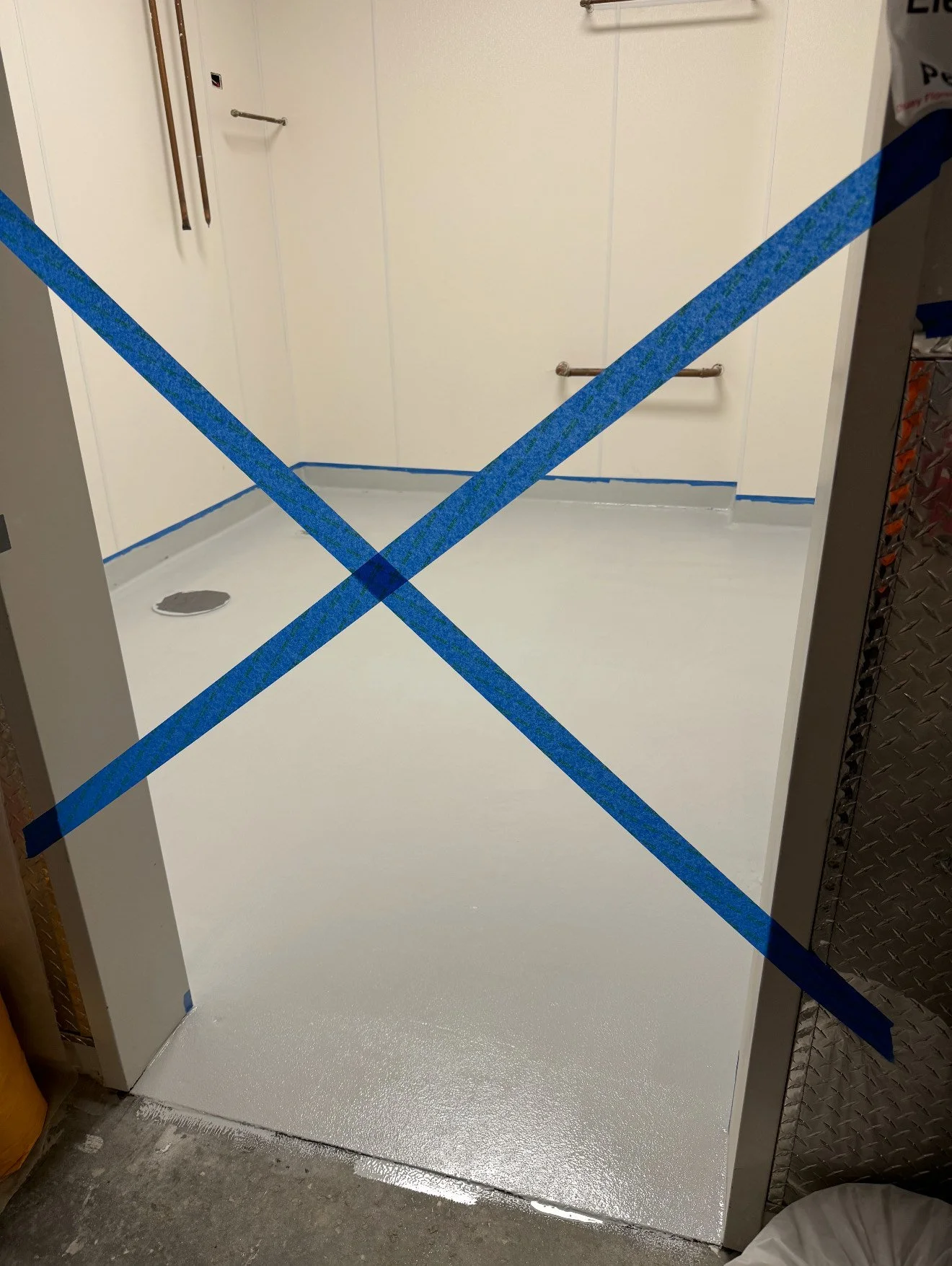 Empty room with white painted walls and a gray floor, blue painter's tape in a large X on the doorway, with a drain in the floor and wall-mounted grab bars, indicating a bathroom under renovation.