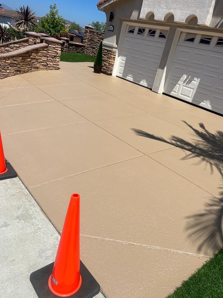 Freshly poured concrete driveway with orange traffic cones, a white garage door, stone wall, small trees, and bright sunny weather.